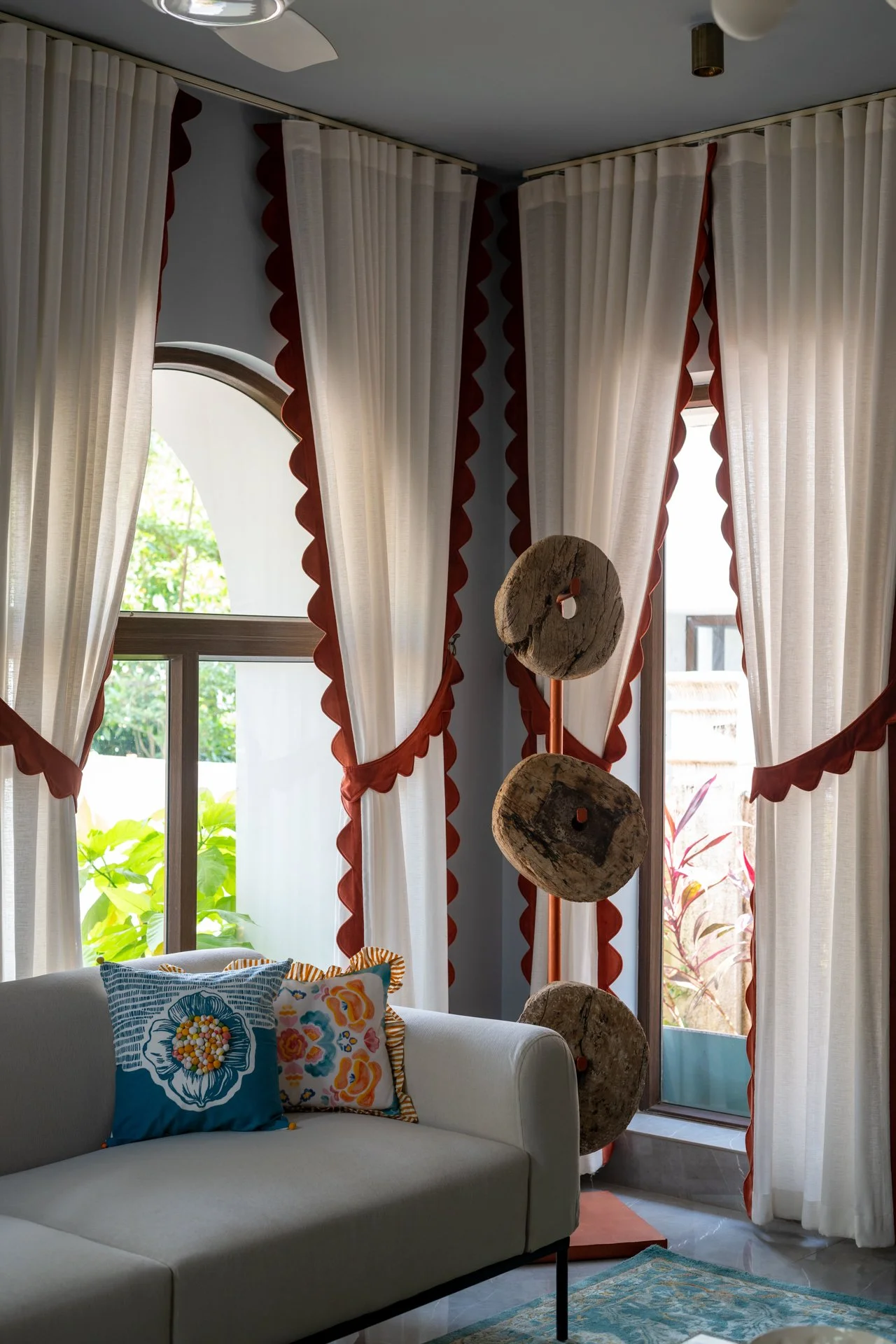 Living room with large white curtains with red scalloped trims, a white sofa with colorful floral pillows, a decorative wood sculpture, and a sliding glass door with a view of greenery outside.
