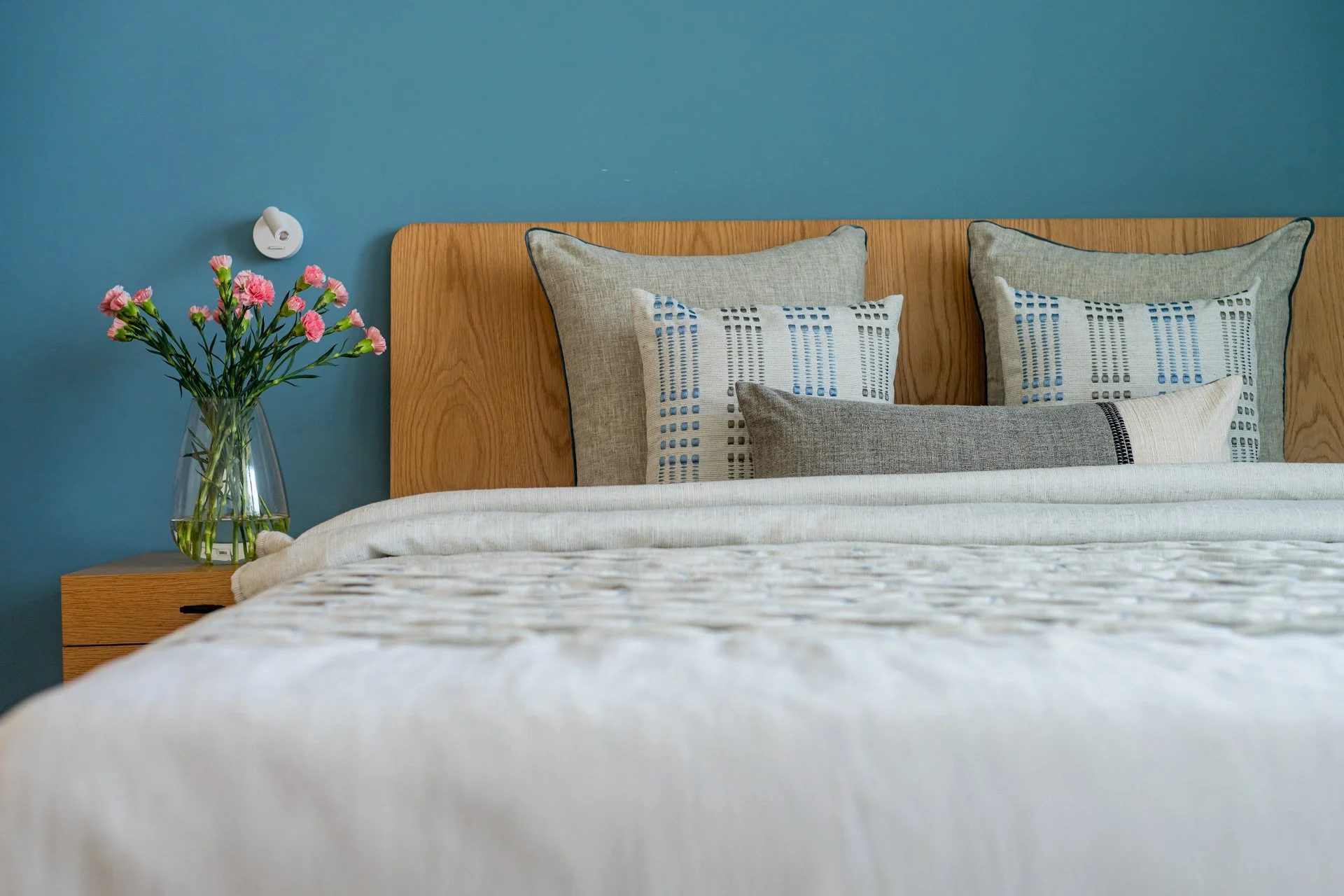 A bedroom with a wooden headboard, beige and patterned pillows, a blue wall, a vase with pink flowers on the bedside table, and a wall-mounted reading light.