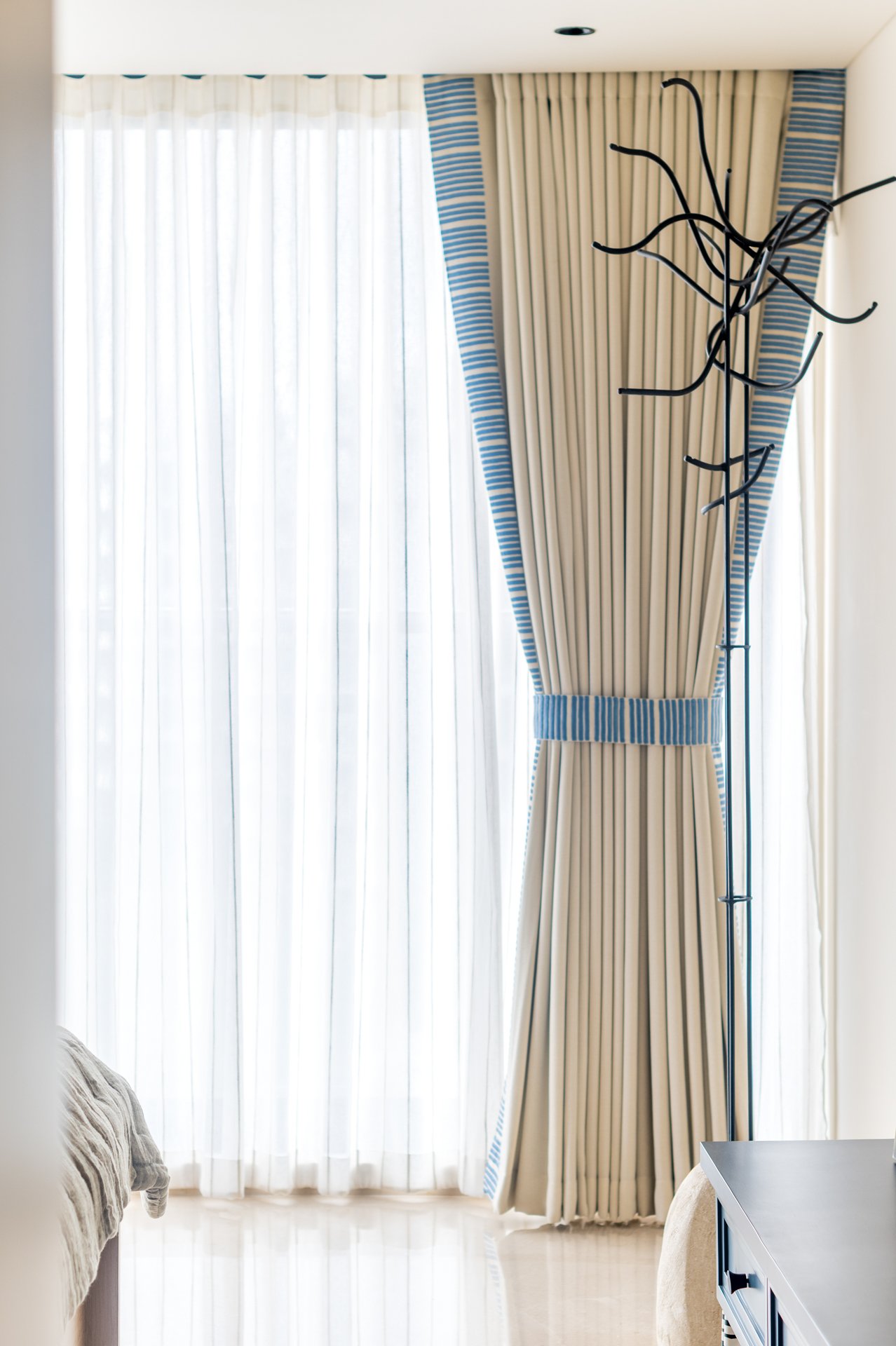 Curtains with sheer white drapes and a striped valance on a large window, with a black abstract floor lamp and partially visible beige upholstered furniture.