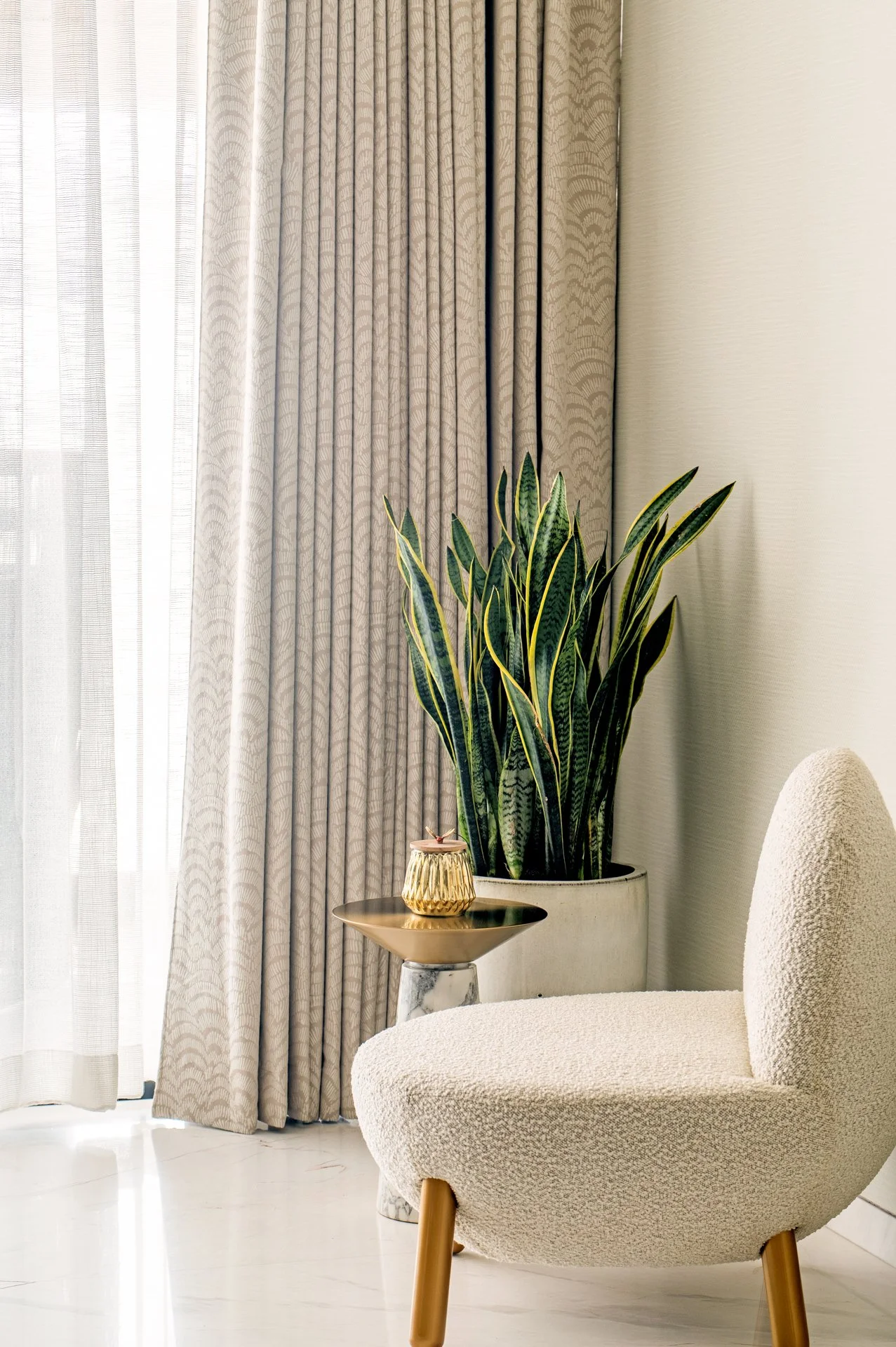A beige upholstered armchair with wooden legs next to a small black marble-topped side table holding a gold decorative object. Behind the chair, there is a large potted snake plant and gold-and-white curtains covering a window or glass door.