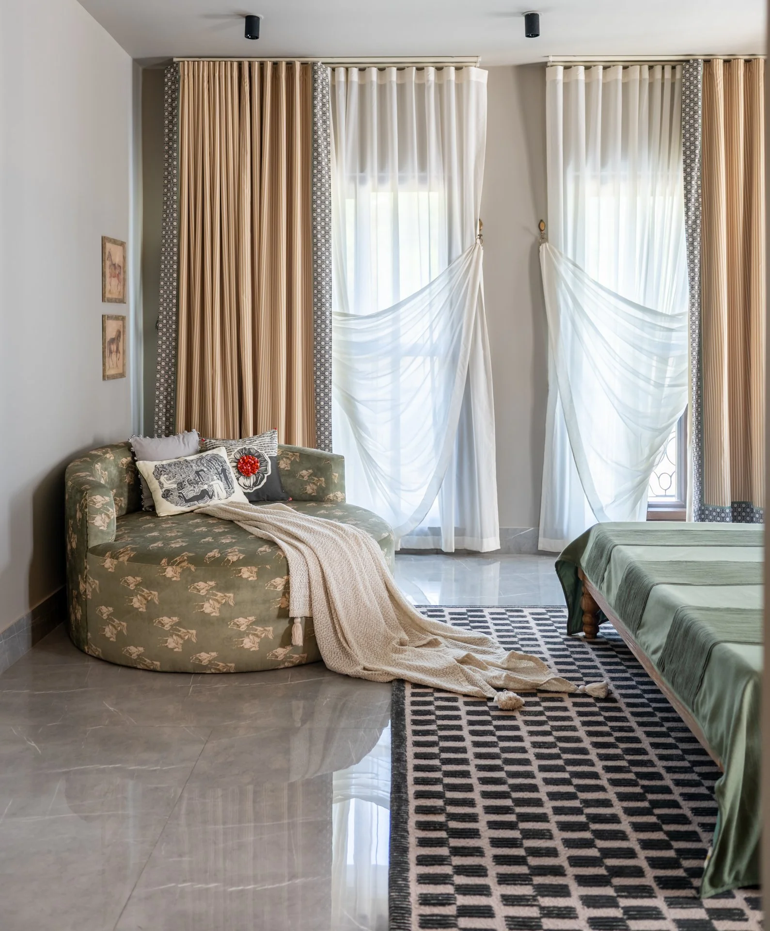 A living room with beige and white curtains, a floral-patterned sofa with pillows, a green-covered bed, and a black-and-white checkered rug on a glossy tiled floor.