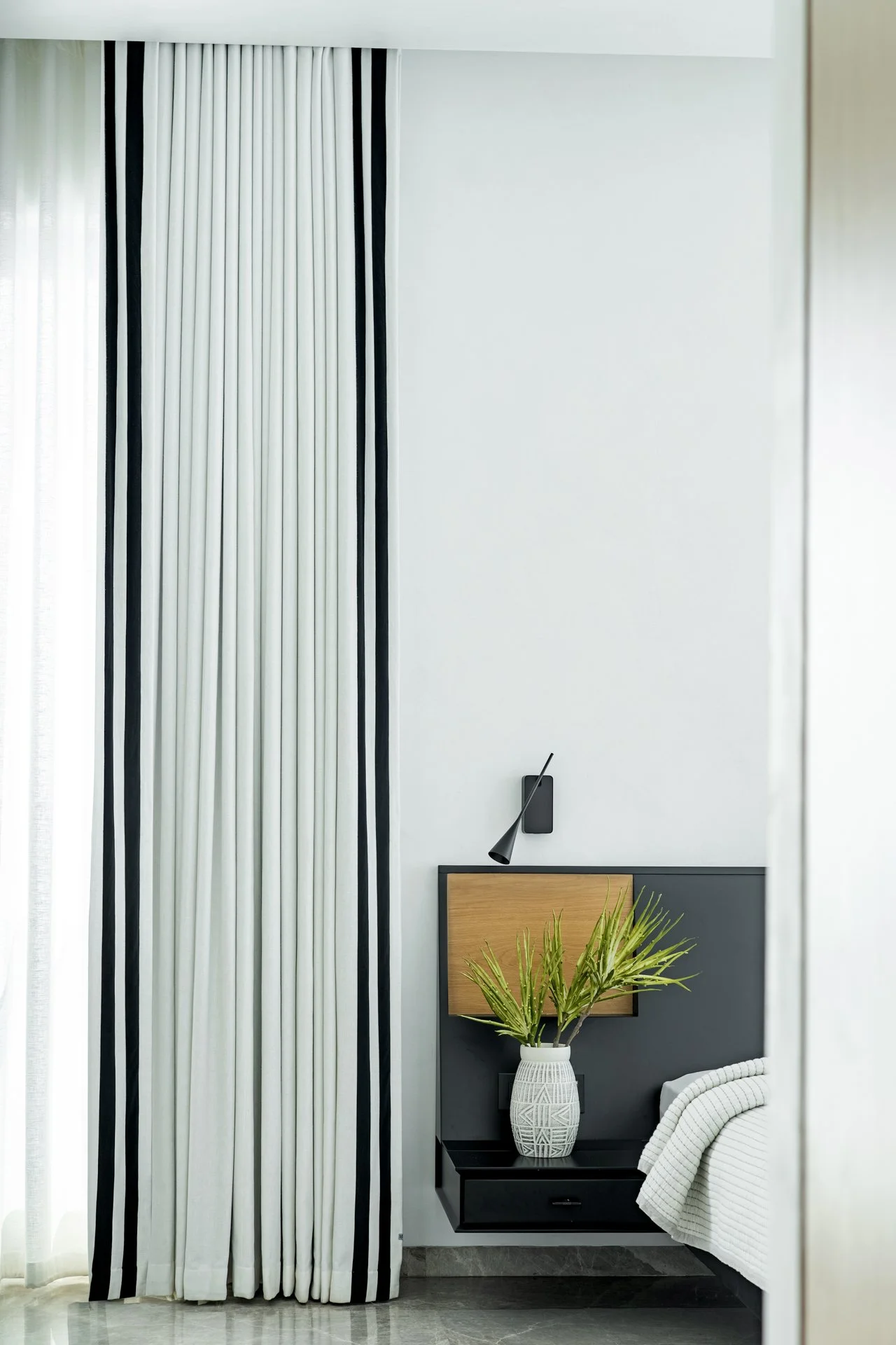 Modern bedroom with black and white color scheme, featuring a curtain with vertical black and white stripes, a wall-mounted black reading lamp, a wooden headboard, a white patterned vase with green palm leaves, and a white bed with a textured blanket.