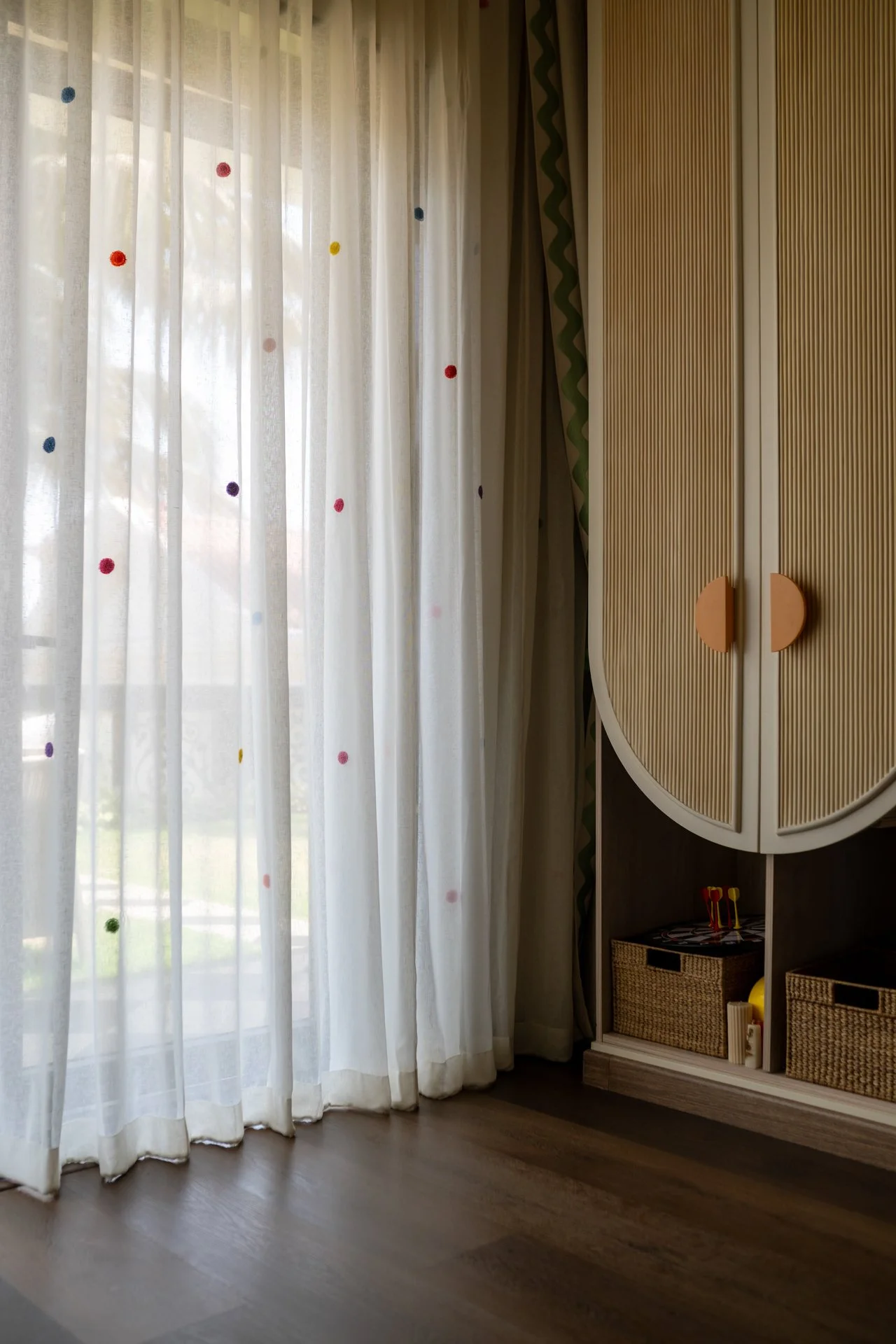 Sheer white curtains with colorful polka dots hanging in front of a window, with a wooden cabinet with rounded doors and orange handles to the right, and a wood floor.