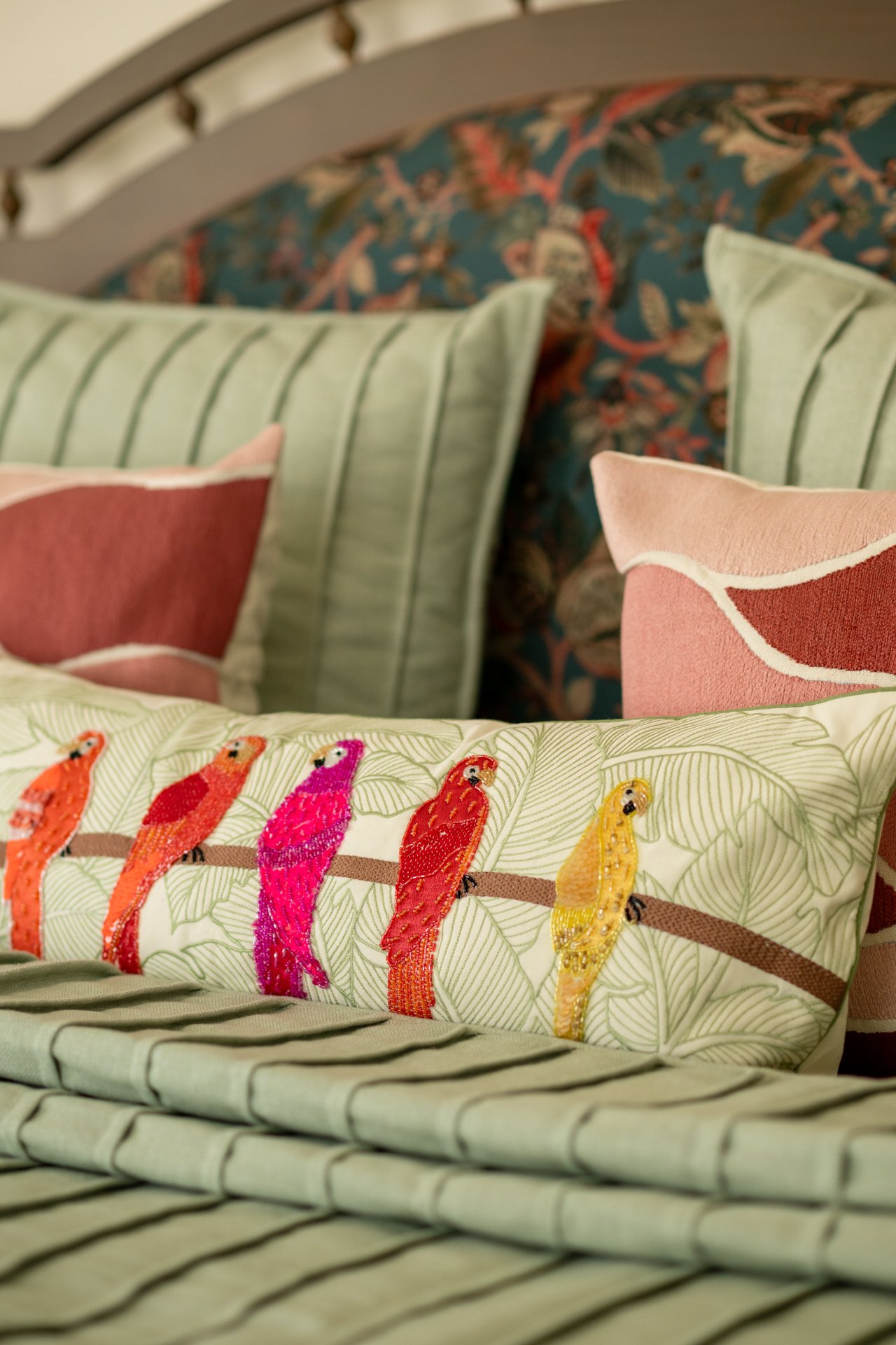 Close-up of decorative pillows on a bed, with one pillow featuring embroidered colorful parrots and lush green leaves.