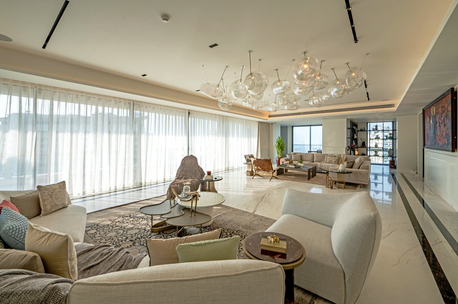 A spacious, modern living room with large windows and sheer curtains, featuring multiple sofas, armchairs, and coffee tables, with decorative lighting fixtures hanging from the ceiling and a bookshelf in the background.