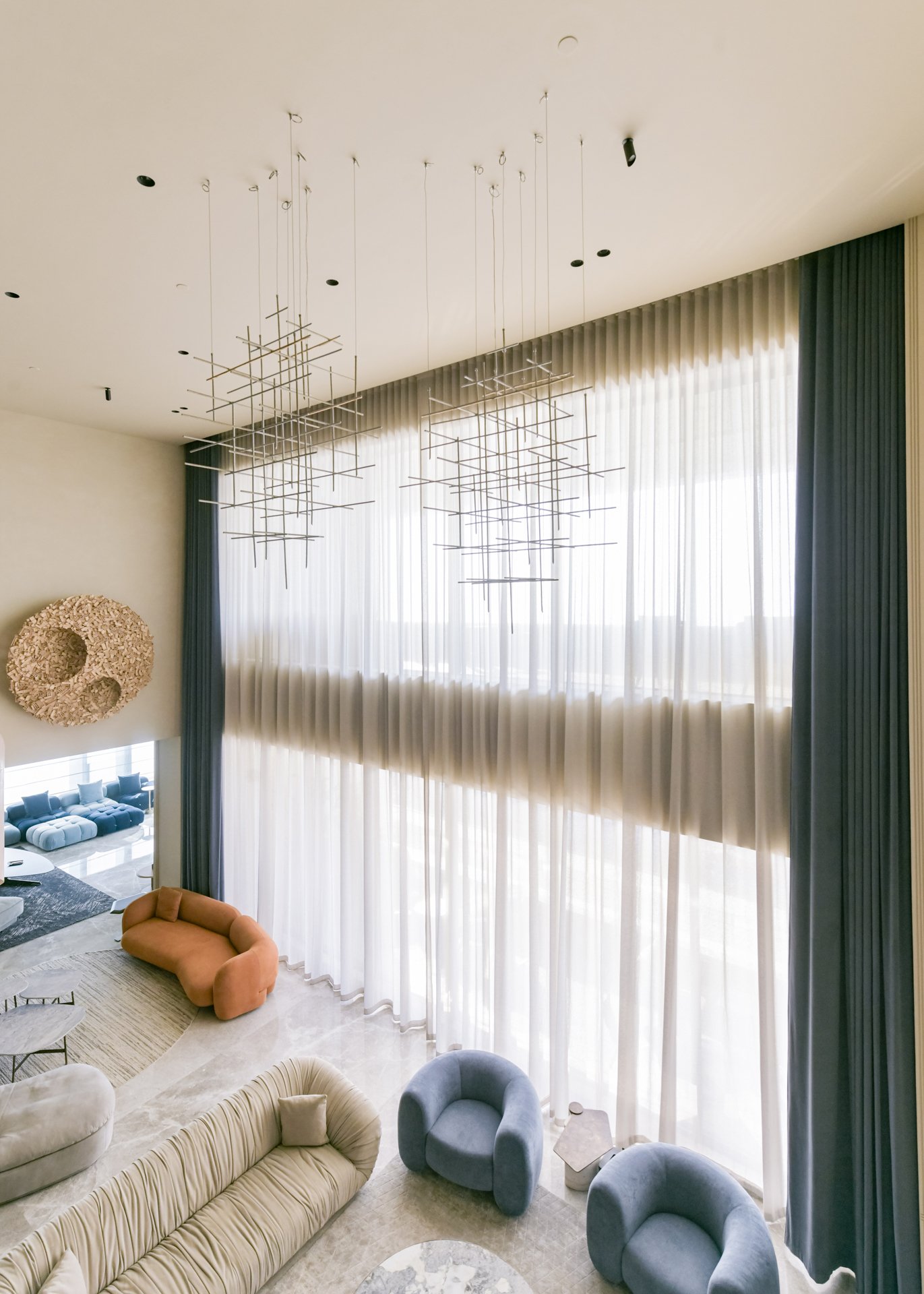 Interior view of a modern, spacious living room with high ceilings, large windows with sheer curtains, and contemporary furniture including lounge chairs and sofas in soft pastel colors. There are also large, decorative light fixtures hanging from the ceiling.