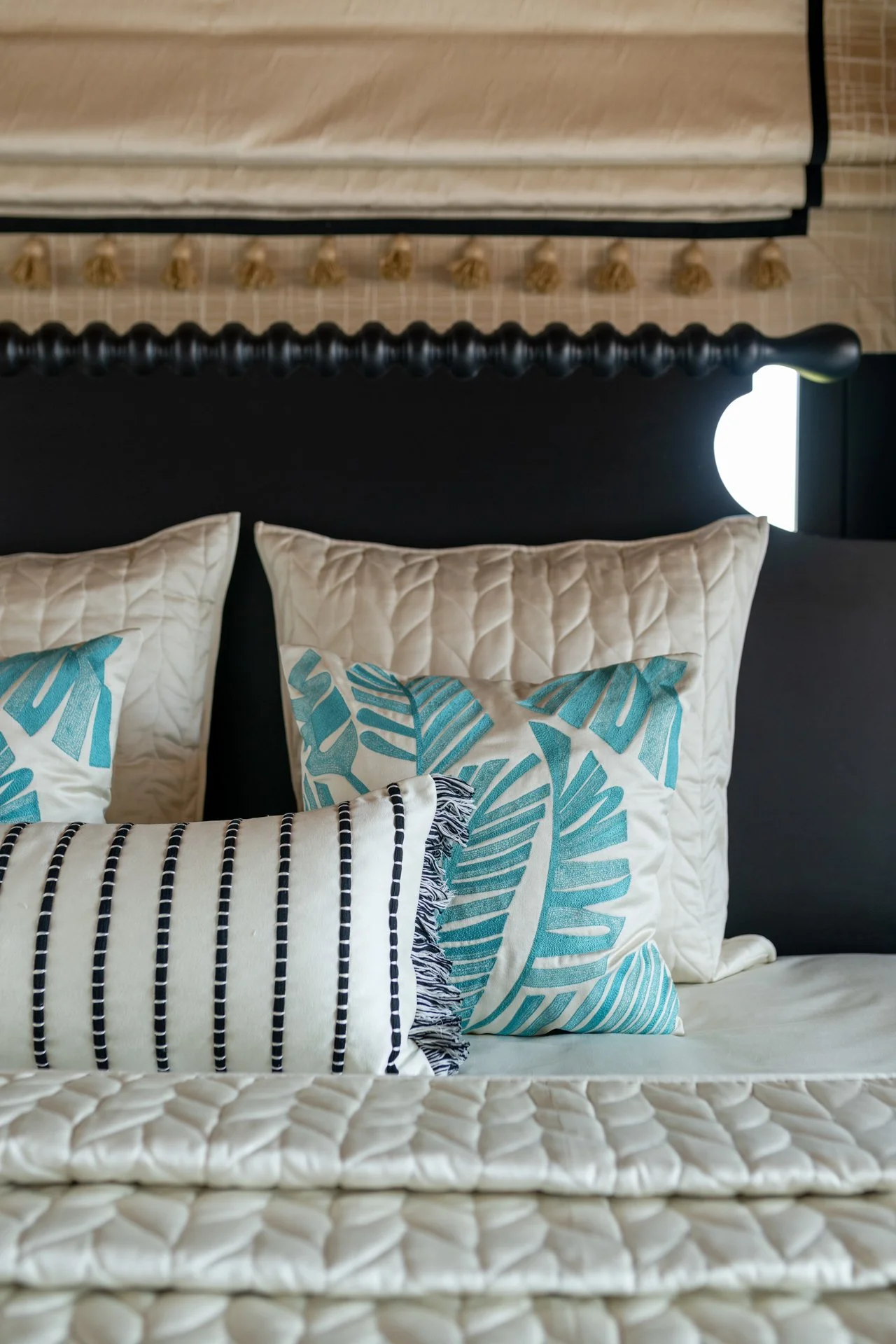 A bed with cream quilted bedding, topped with multiple decorative pillows, including one with blue tropical leaf patterns and another with black and white stripes, against a black headboard with large pillows, in a bedroom with a modern, minimalist design.