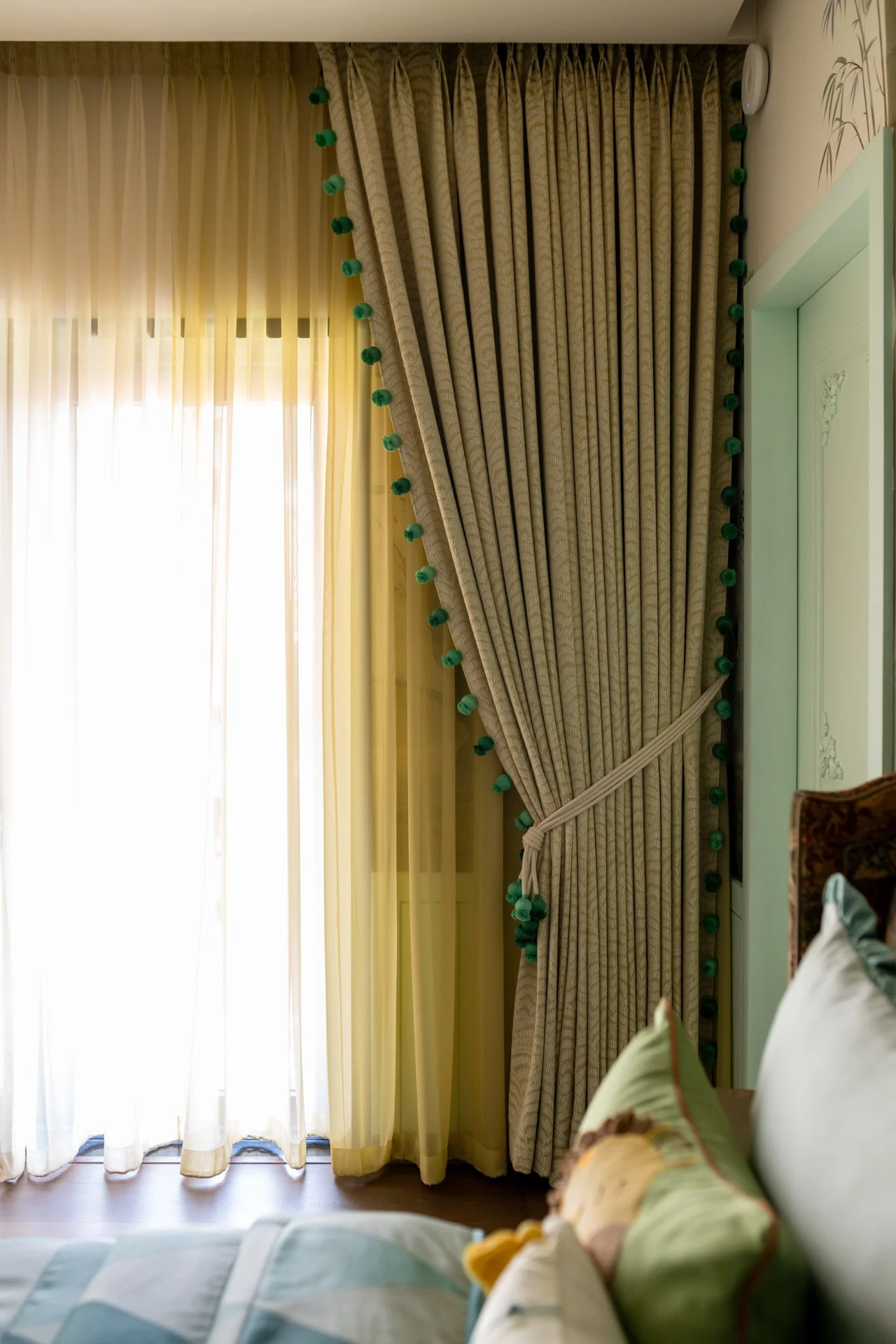 Sunlight coming through white sheer curtains and beige drapes with green pom-poms on the curtain tie-back in a bedroom.