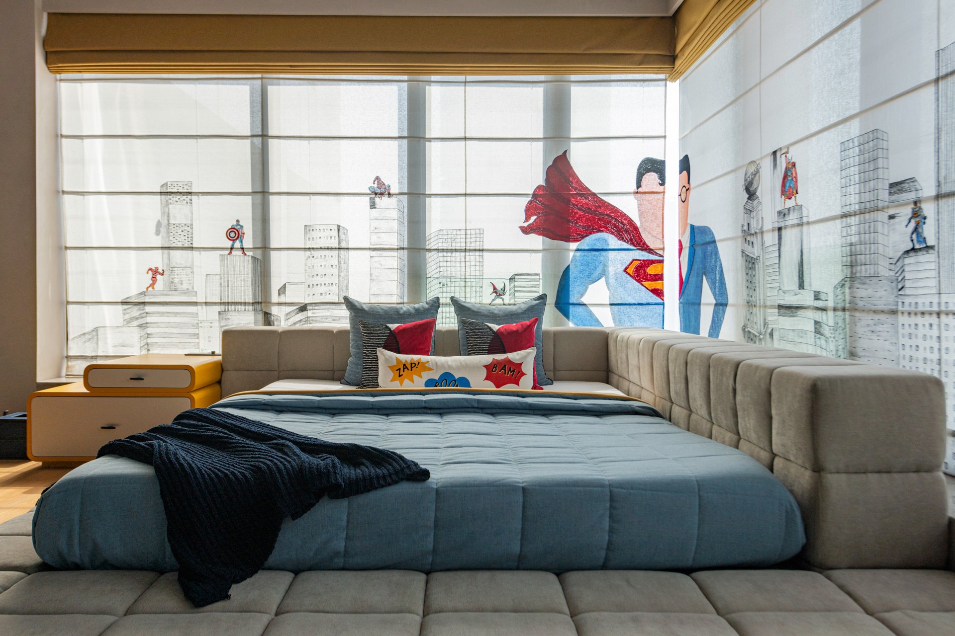 A modern hotel room with a large bed and a cityscape mural featuring superhero imagery on the window covering. The bed has a blue comforter, black throw, and comic-style pillows. There is a beige tufted seating area around the bed.