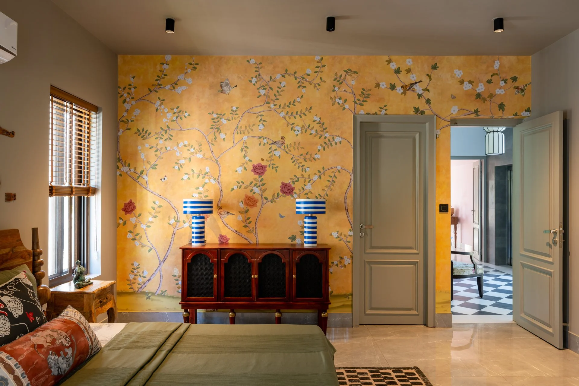 Interior view of a room with a yellow wall featuring a painted floral and bird mural, a wooden sideboard with striped blue and white lamps, a window with blinds, and an open door leading to a hallway with black and white checkered flooring.