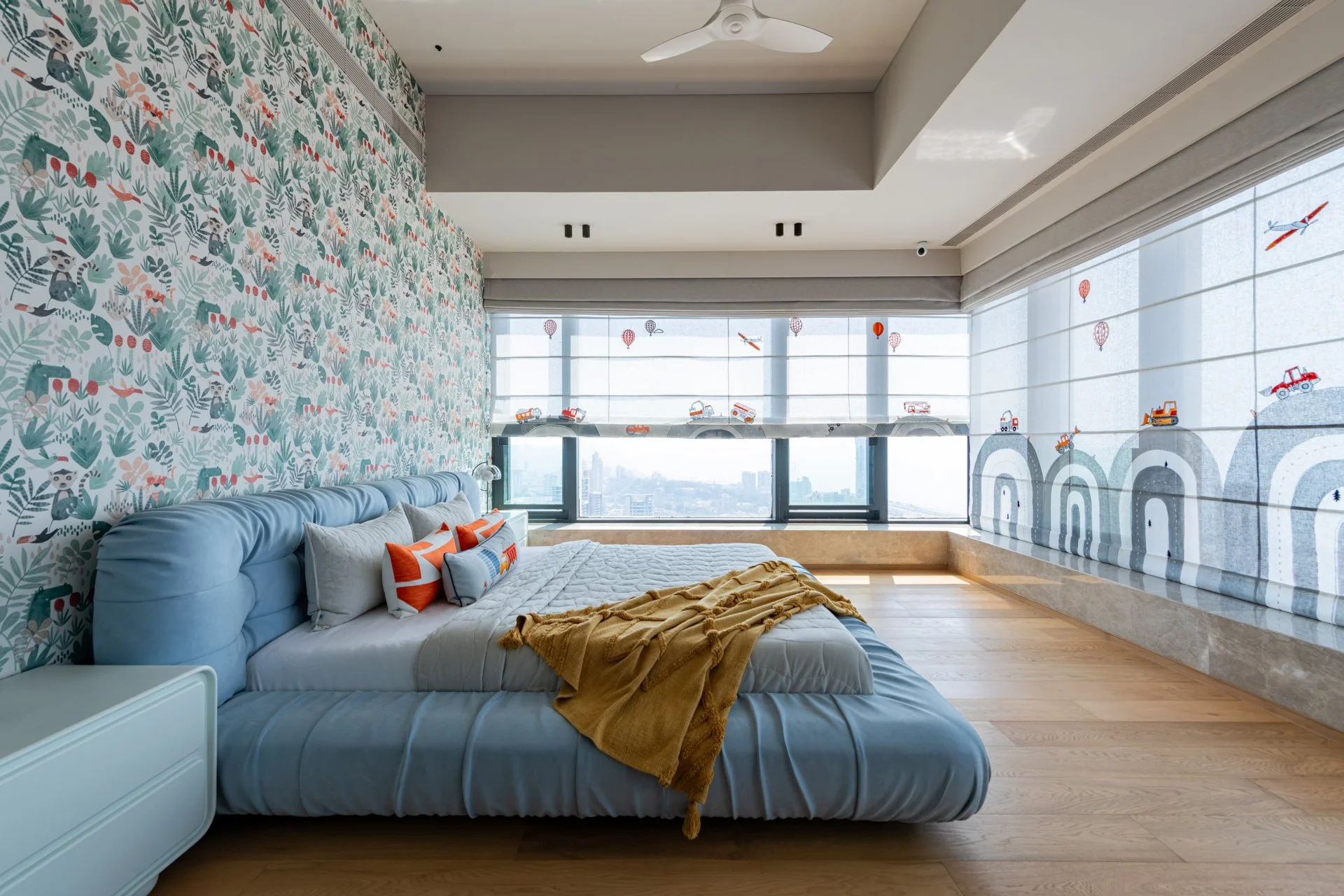 A modern bedroom with light blue upholstered bed, mustard yellow blanket, white side table, decorative pillows, patterned wallpaper with animals and plants, and large windows with blinds featuring construction vehicles and hot air balloons.