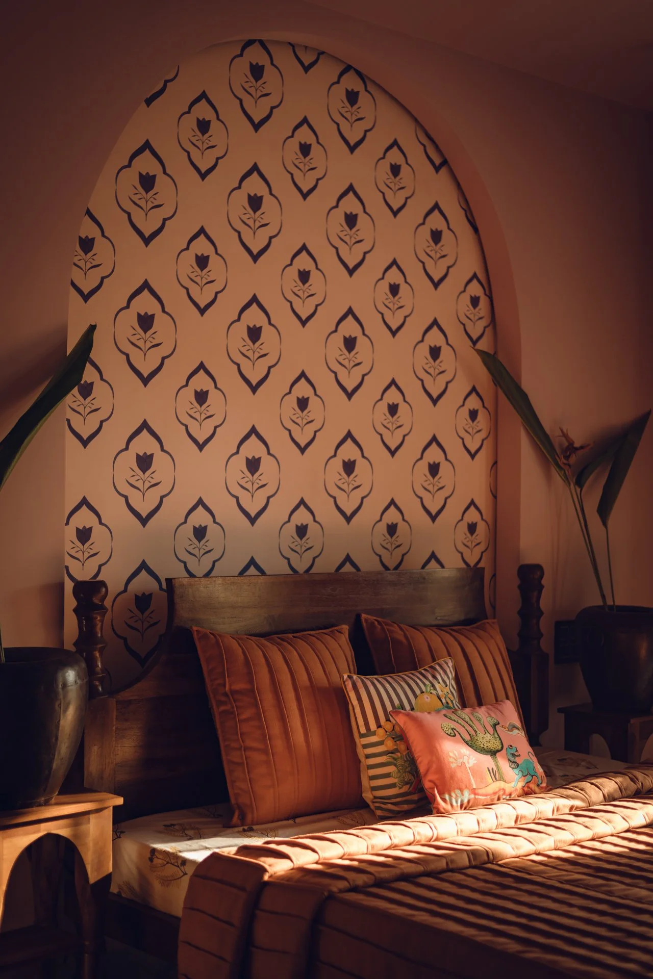 A cozy bedroom with an arched wall covered in patterned wallpaper featuring dark navy floral motifs on a beige background. The bed has a wooden headboard, rust-colored pillows, and a decorative pillow with colorful animal illustrations. There is a bedside table with a large dark vase, and houseplants are on either side of the bed.