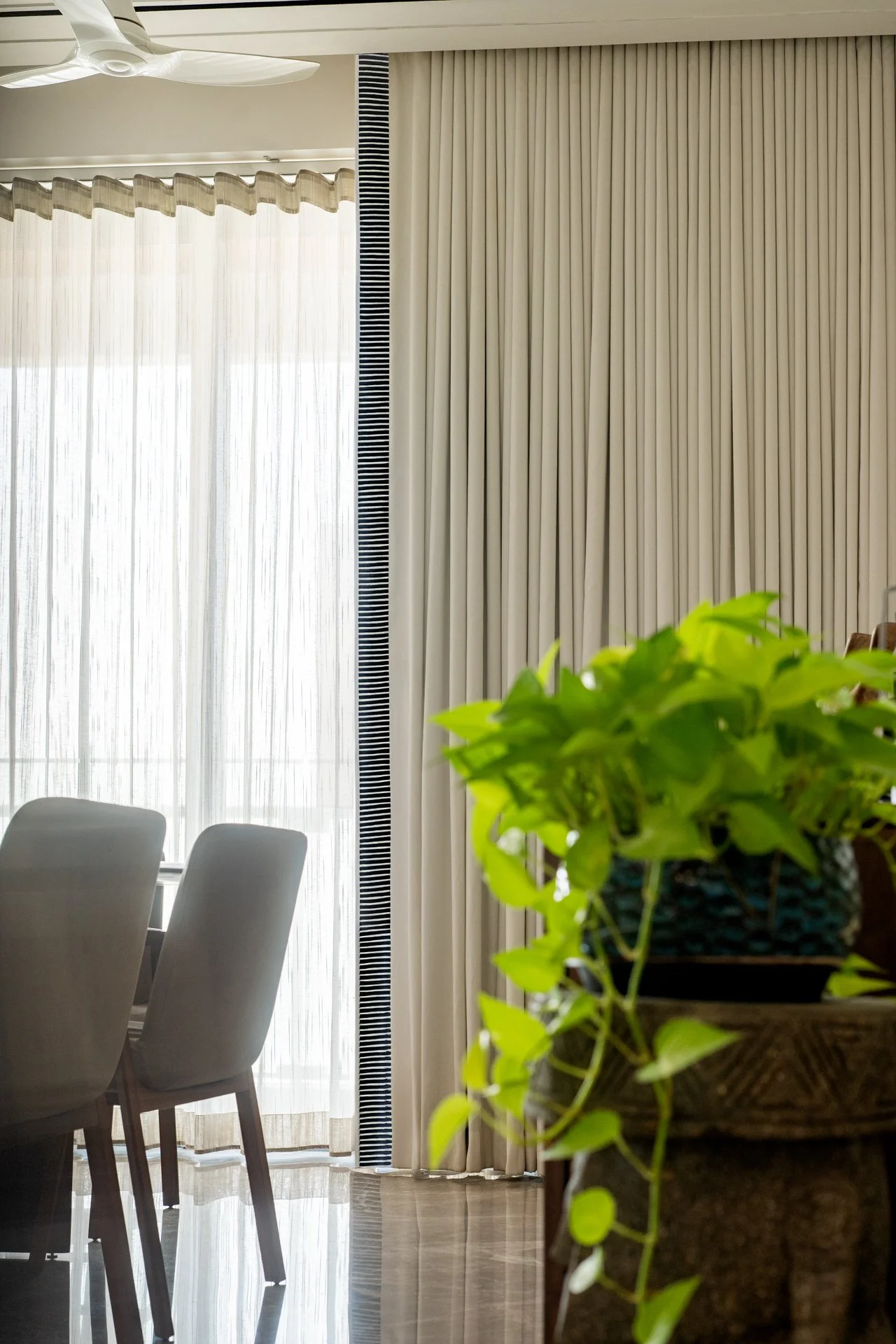 A dining room with white chairs, sheer white curtains, and cream-colored blackout curtains, with a houseplant in the foreground.