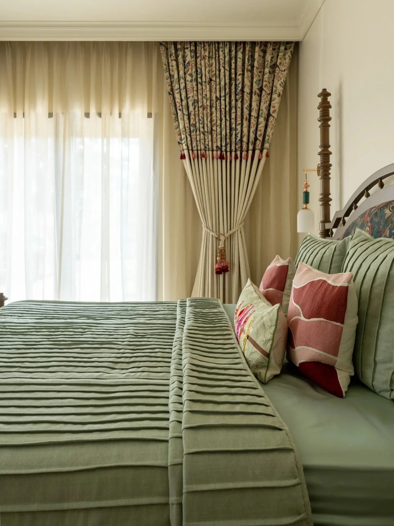 A bedroom corner with a bed, green-striped bedding, and decorative pillows. There are heavy floral-patterned curtains tied back, allowing light into the room.