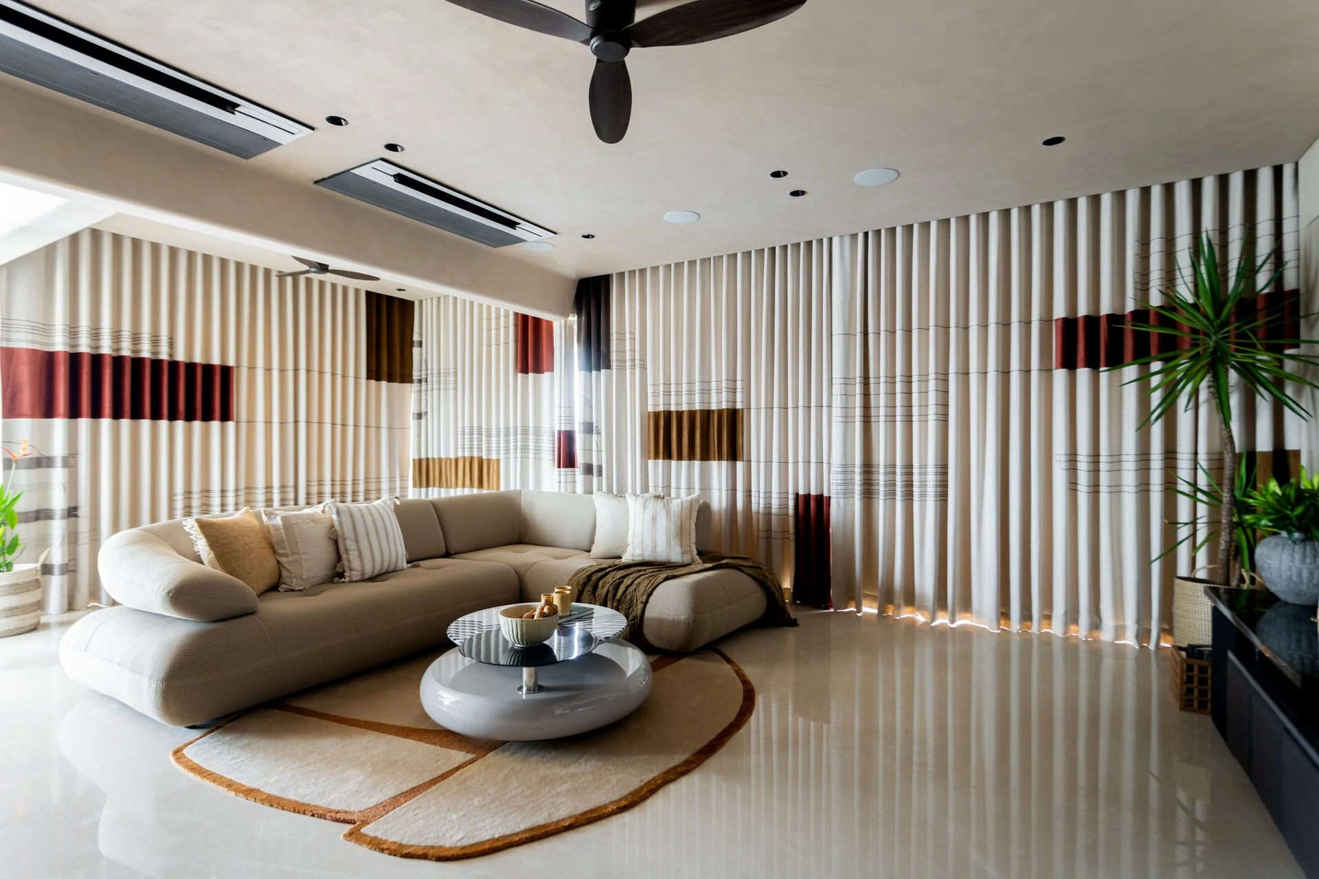 A modern living room with beige curtains, a light-colored sofa with pillows, a round coffee table with bowls, a decorative rug, a ceiling fan, and potted plants, with recessed lighting on the ceiling.