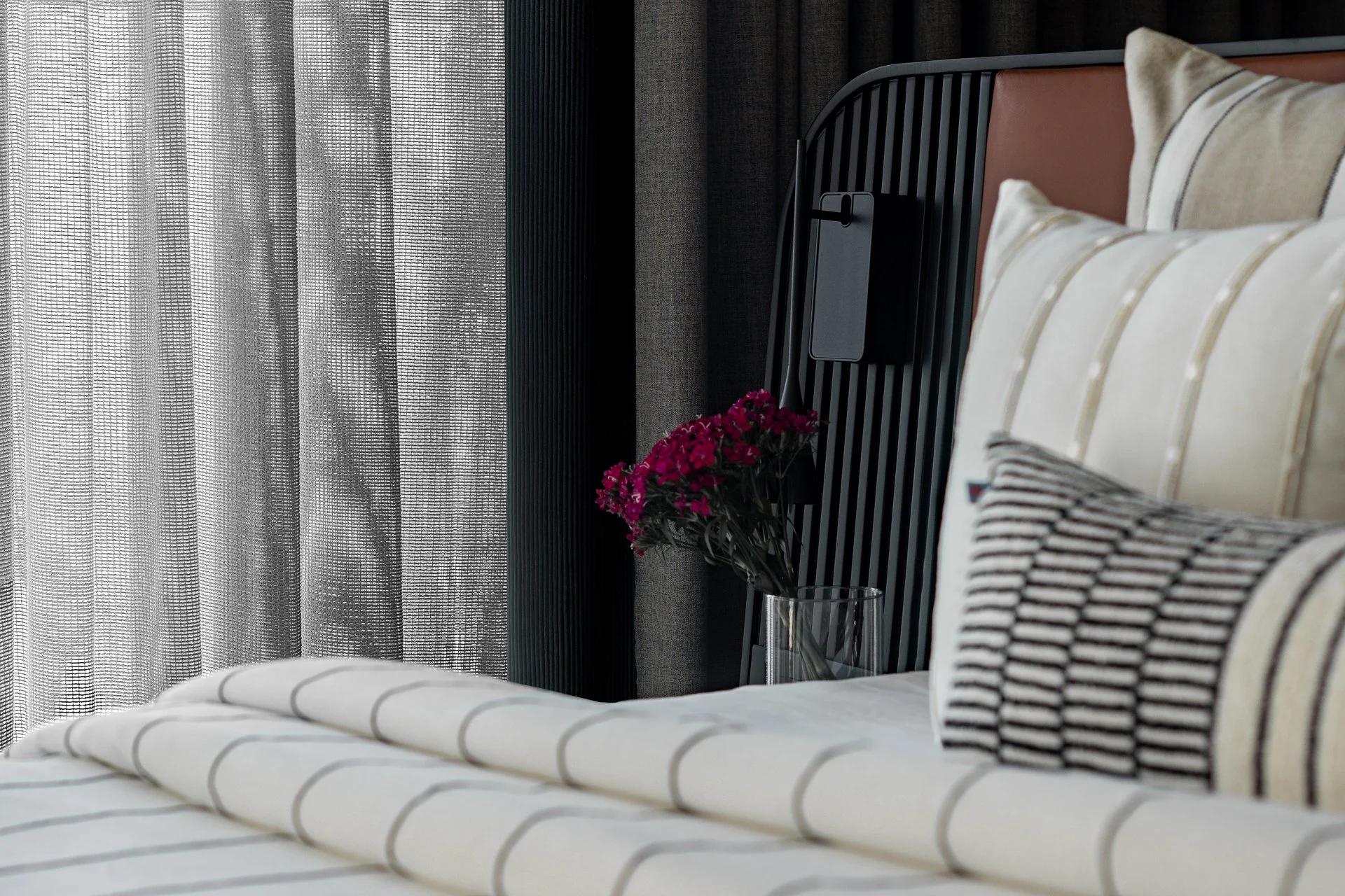 Close-up of a bed with white and gray striped bedding, pillows, a side table with a small vase of pink flowers, behind which are dark curtains, and a section of a headboard.
