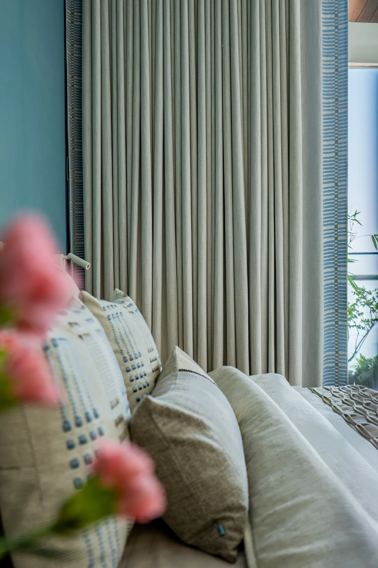 Living room with beige couch and pillows, beige curtains, and a window with plants outside.