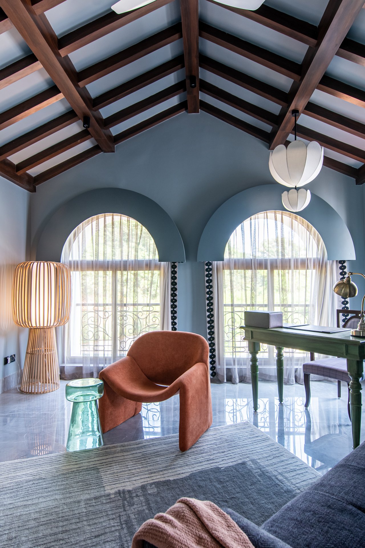 A cozy living room with a high ceiling featuring wooden beams. Large arched windows with sheer curtains let in natural light. Decor includes a rust-colored armchair, a green side table, a tall wooden floor lamp, and a desk with a greenish finish and a brass desk lamp.