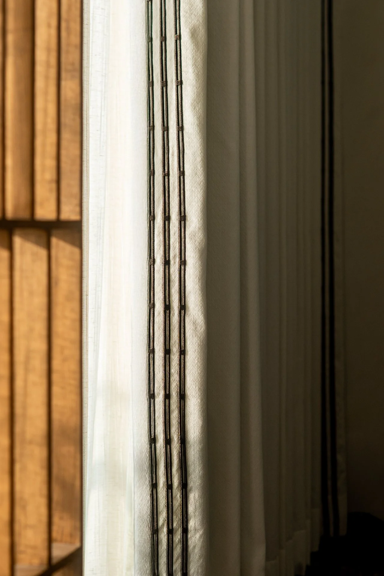 Close-up of a window with vertical blinds, with sunlight filtering through. The blinds are partially open, revealing a sheer curtain behind them.