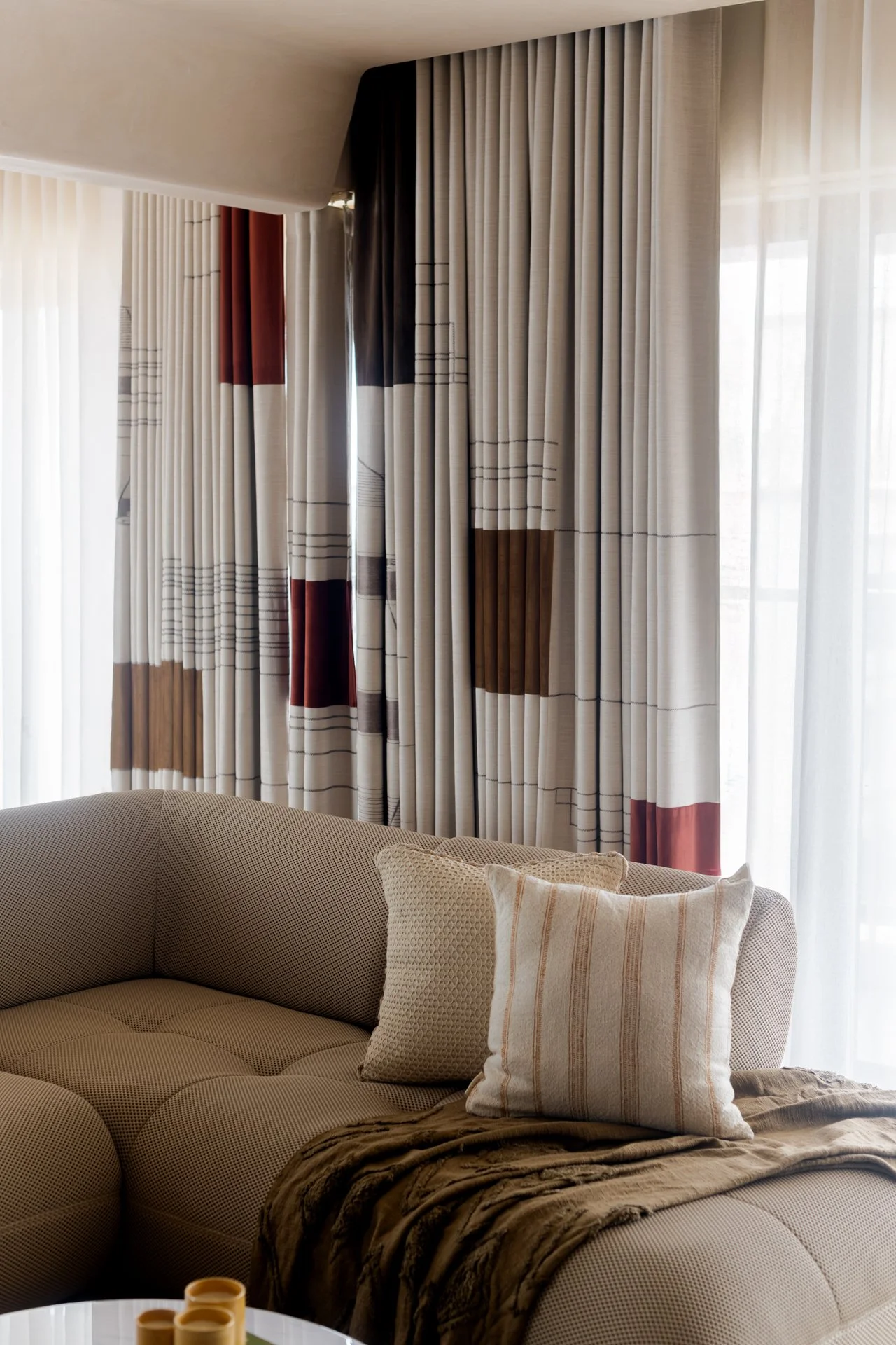 Living room with beige couch, patterned throw pillows, and layered drapes in neutral and brown tones, with sunlight filtering through.