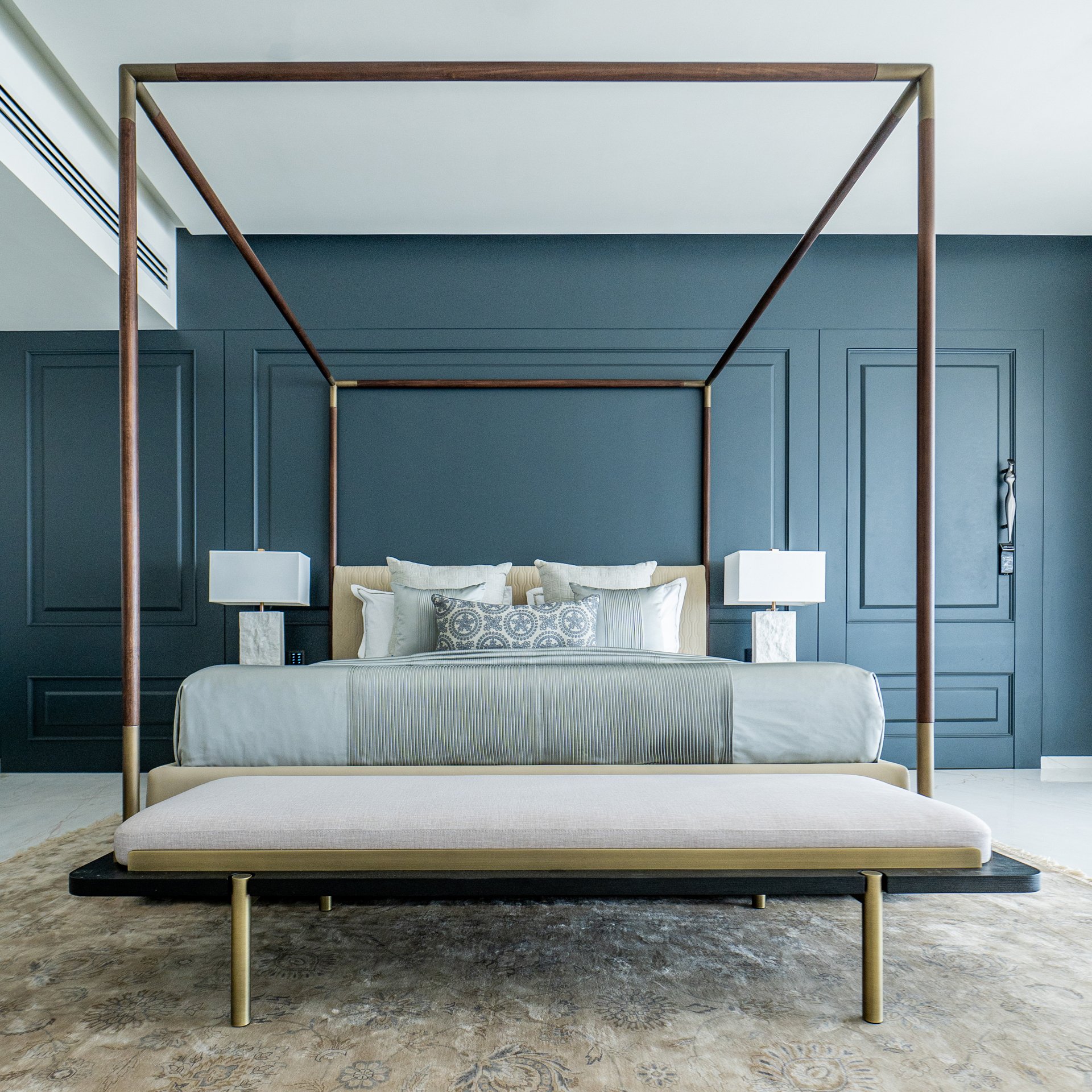 Luxurious bedroom with a modern bed featuring white textured bedding and multiple pillows, a dark blue accent wall with decorative paneling, and a four-poster frame made of wood and metal, standing on a marble floor.