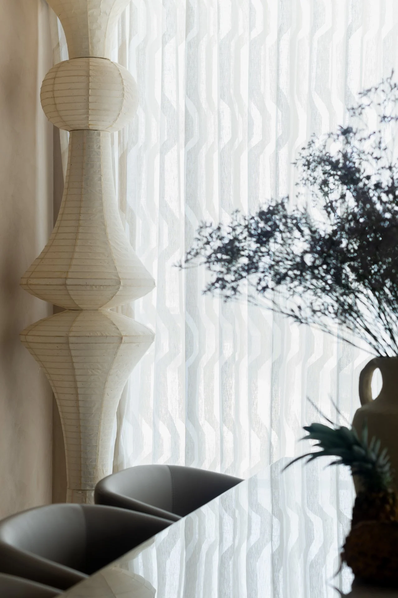 Sunlit modern dining room with white paper lantern-style vase or sculpture, patterned curtains, and potted plant on table, viewed through window.