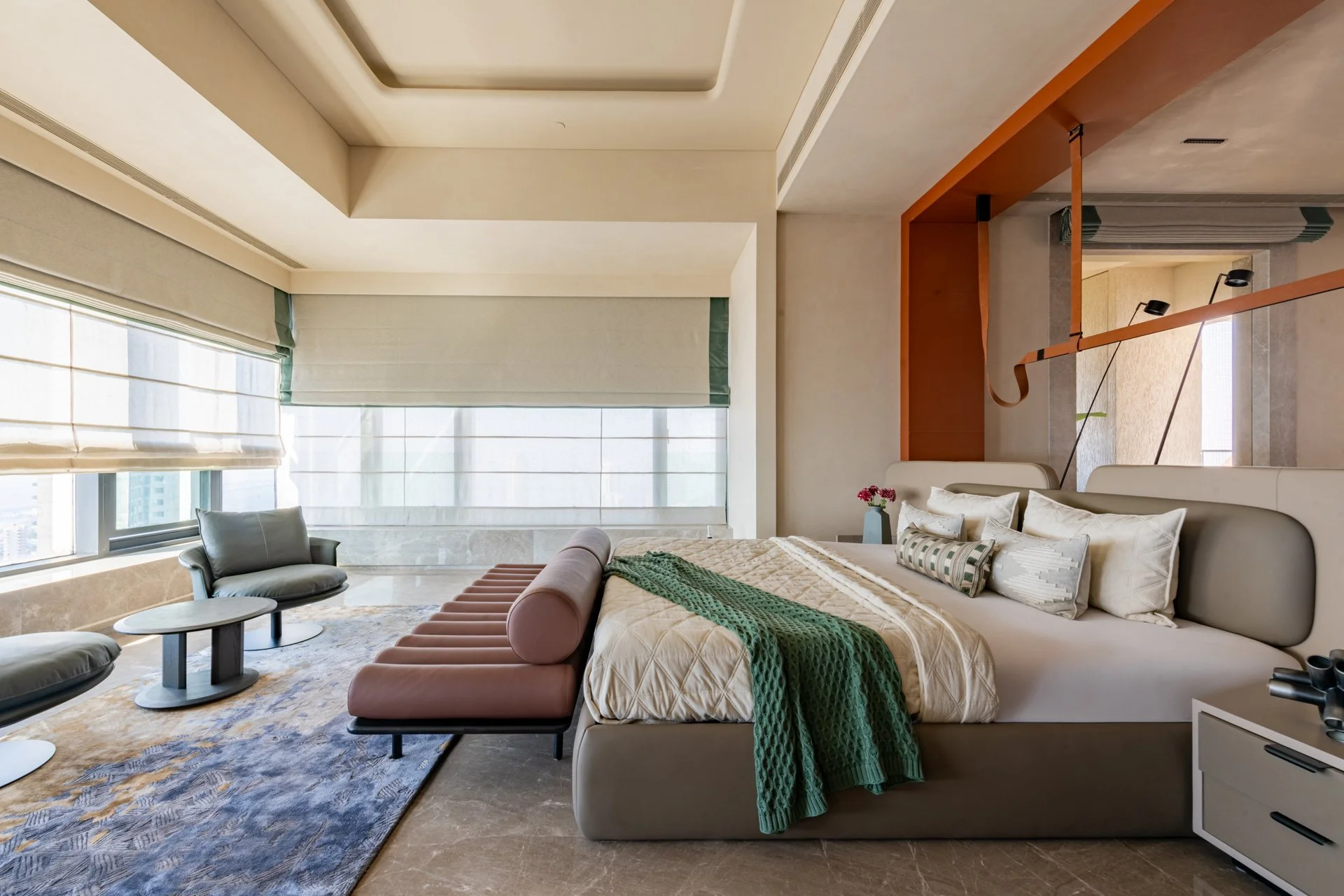 Modern hotel room furnished with a large bed with multiple pillows, a green blanket, and a beige bedspread. There is a beige sofa at the foot of the bed. Two black chairs and two small tables are near large windows with shades, allowing natural light into the room. A decorative vase with red and pink flowers is on a side table. The ceiling features a recessed design, and there is a wooden curtain rod with attached lights over a seating area.