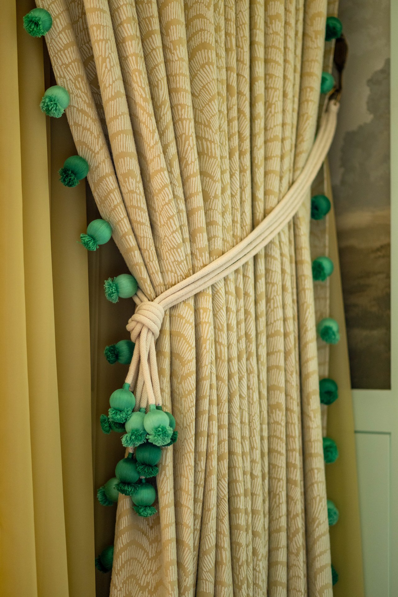 Decorative curtains with a yellow pattern and green pom-pom trim tied back with a yellow cord.