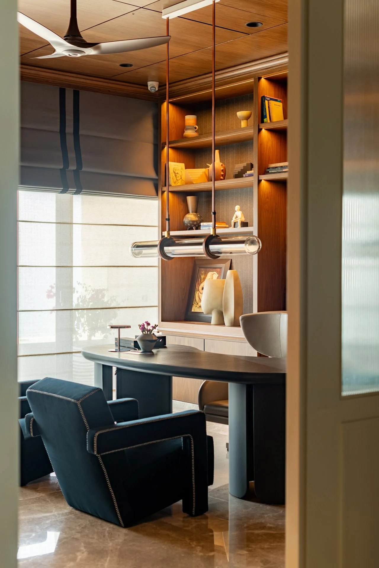 A modern home office or study room with a dark armchair, a round table with a small flower arrangement, built-in wooden shelves with decorative items and books, a ceiling fan, large windows with shades, and a marble floor.