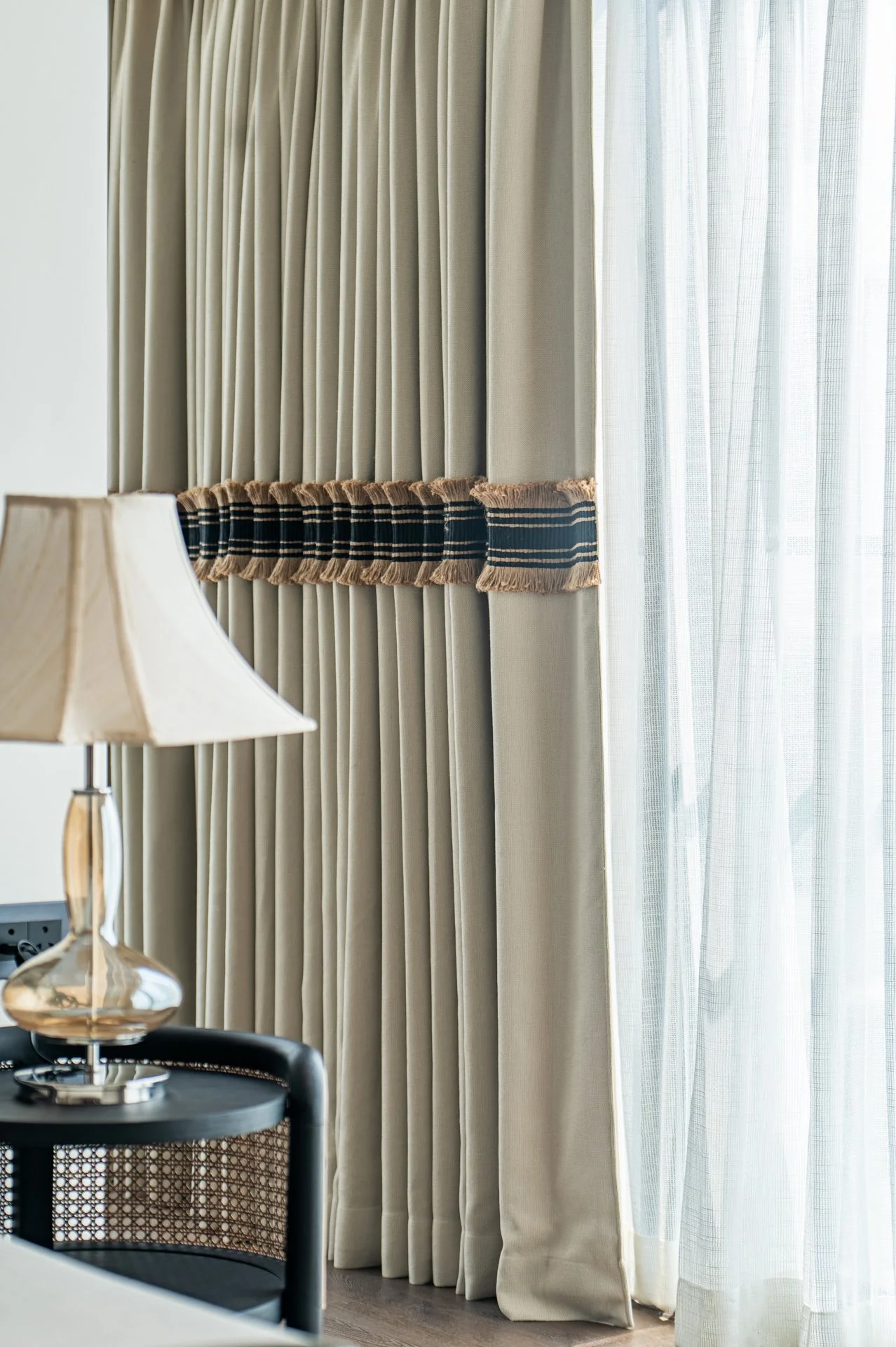 Close-up of beige curtains with a decorative band, partially covering a sheer curtain, in a well-lit room.