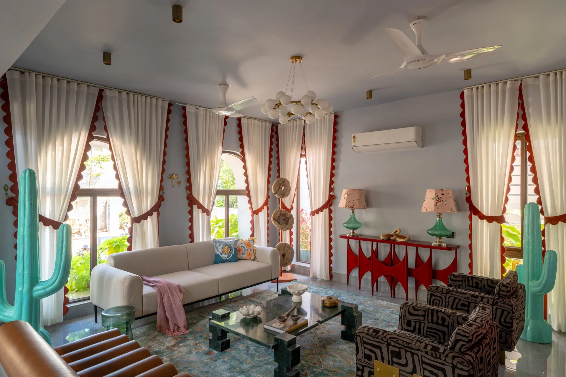 Living room with sheer curtains edged in red, white sofa with colorful pillows, patterned armchair, glass coffee table with decorative items, large cactus sculptures, and a sideboard with lamps, all under a ceiling with fans and a chandelier.