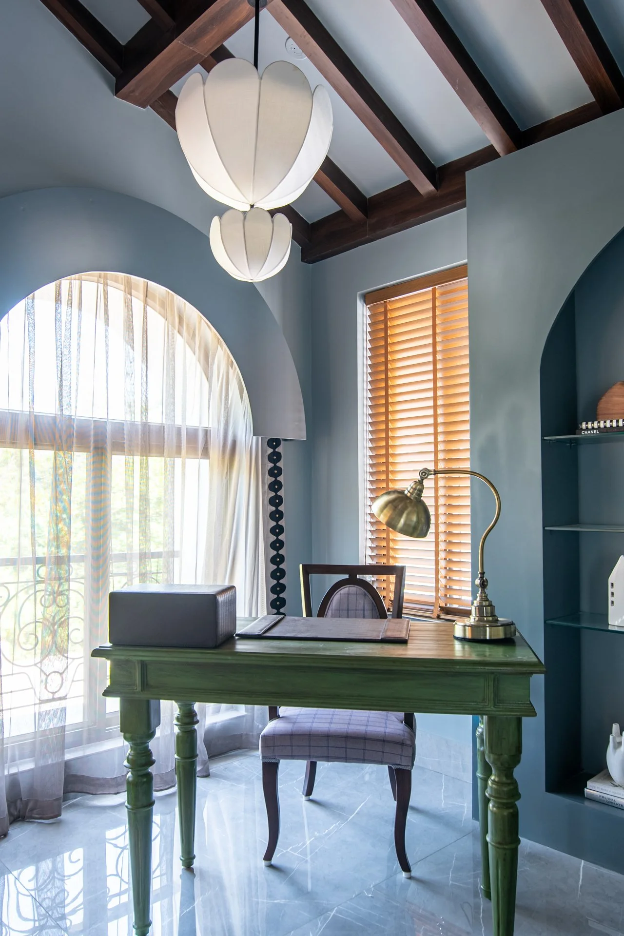 A cozy home office with a wooden desk, a plaid upholstered chair, a brass desk lamp, and large windows with wooden blinds and sheer curtains, illuminated by a white petal-shaped hanging lamp.