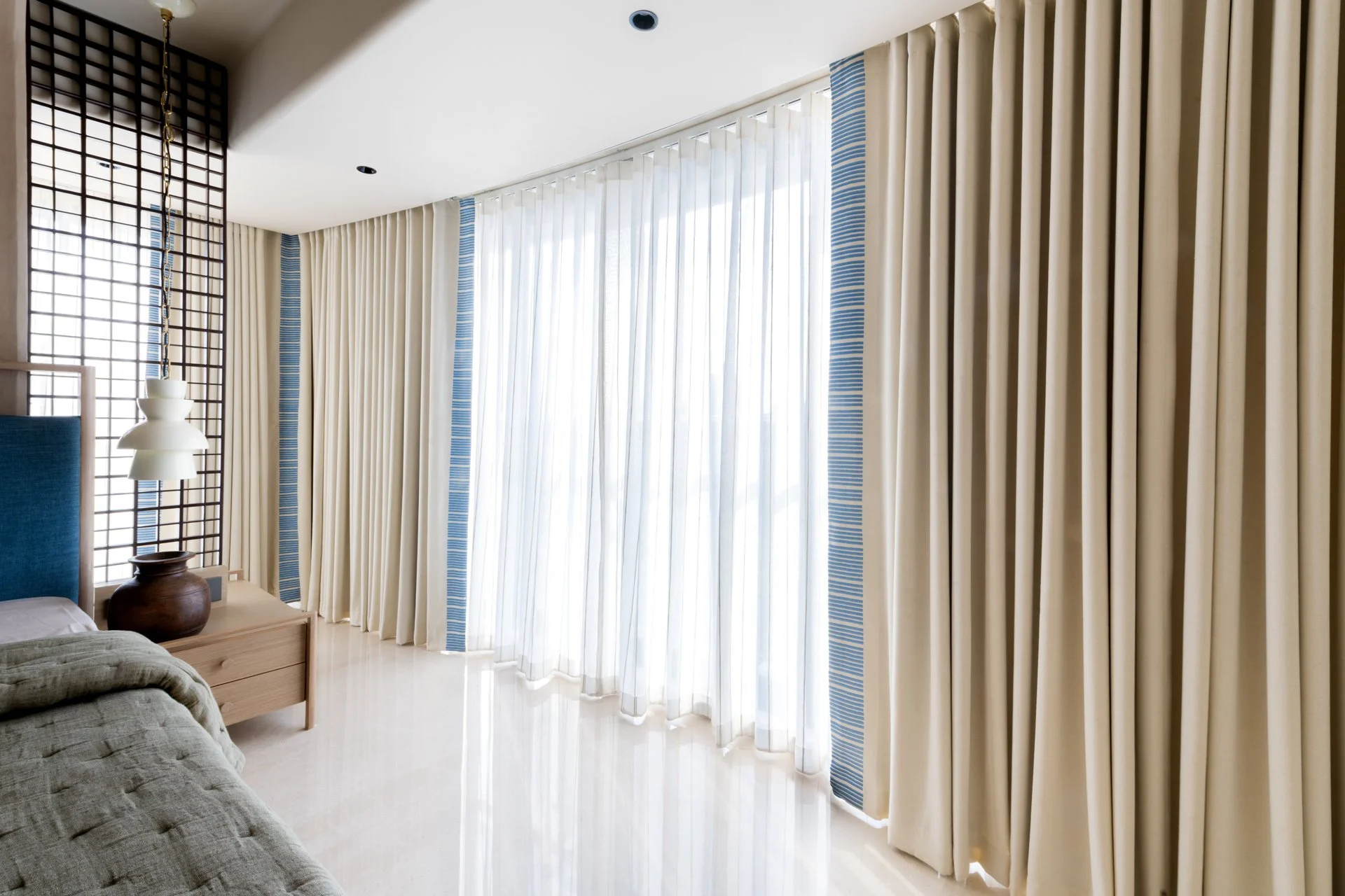 Living room with floor-to-ceiling curtains, a wooden side table with a decorative vase, a large window covered with sheer white curtains, and a couch with a green throw blanket.