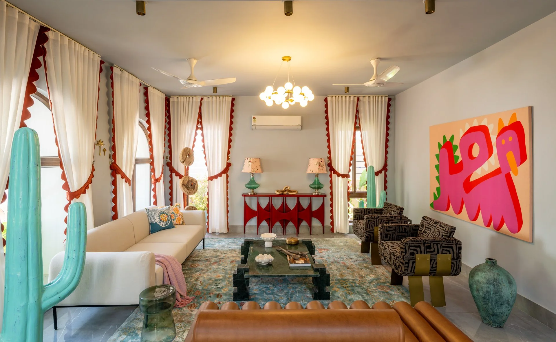 Living room with white sofa, checkered armchairs, colorful artwork on the wall, large windows with white curtains and red trim, decorative lamps, ceiling fans, and desert-themed decor including large cactus sculptures and hanging orbs.