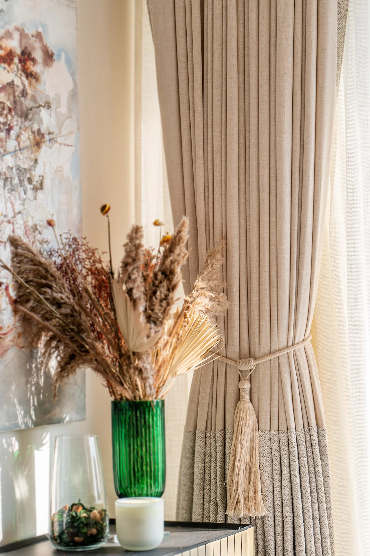 Interior decor with dried flowers in a green vase, a fresh candle, and an abstract artwork on the wall near beige curtains.