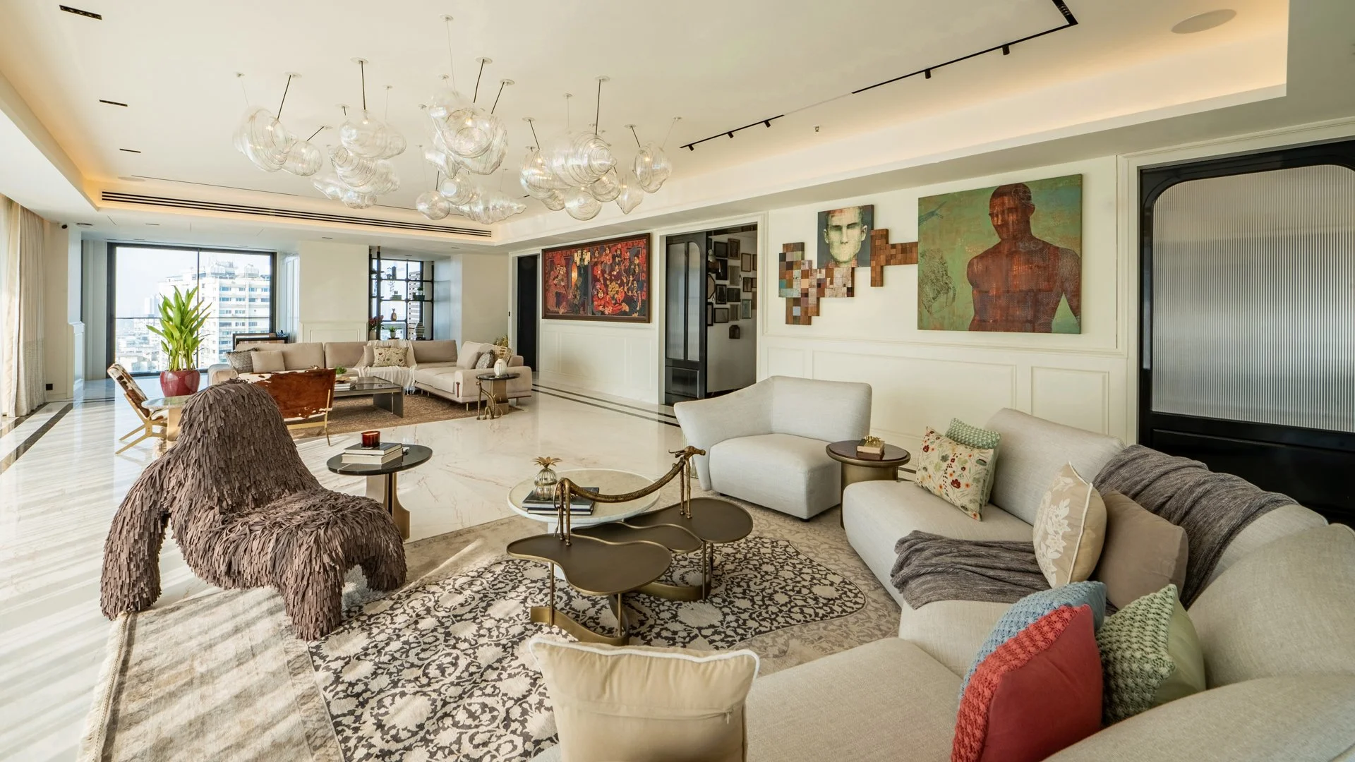 A spacious living room with cream-colored sofas, modern art on the walls, large windows with city views, and decorative lamps hanging from the ceiling.
