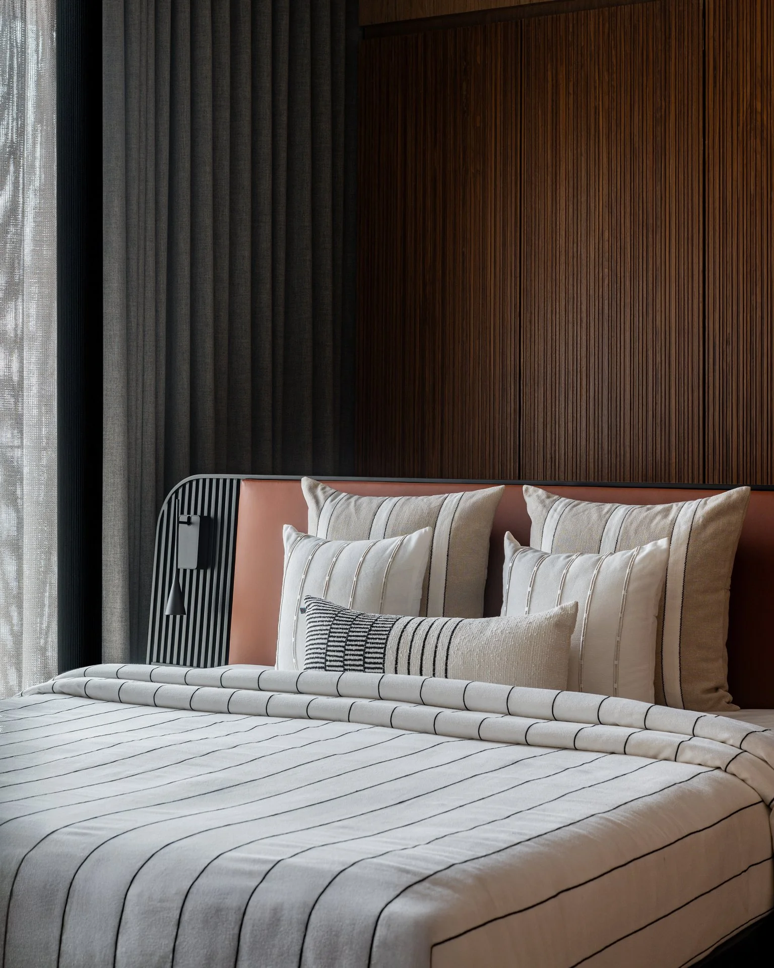 A neatly made bed with a white duvet featuring thin black stripes, multiple decorative pillows with various striped patterns, and a pink headboard. Behind the bed is a dark wood textured wall, and on the left side, a large window with sheer curtains.