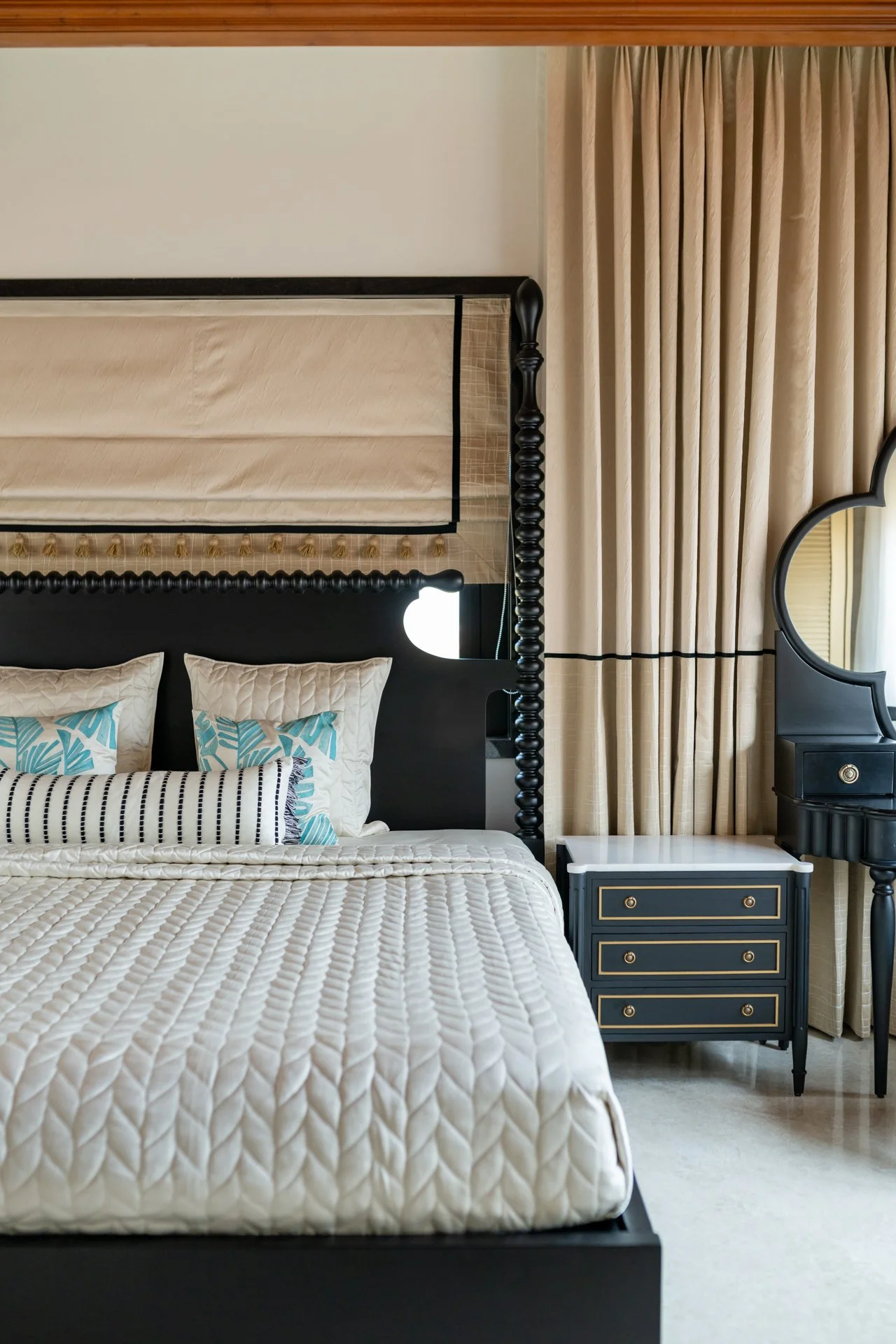 Bedroom with a bed featuring a black headboard and decorative pillows, beige curtains, a black and gold nightstand, and a black mirror with a tall, curved frame.