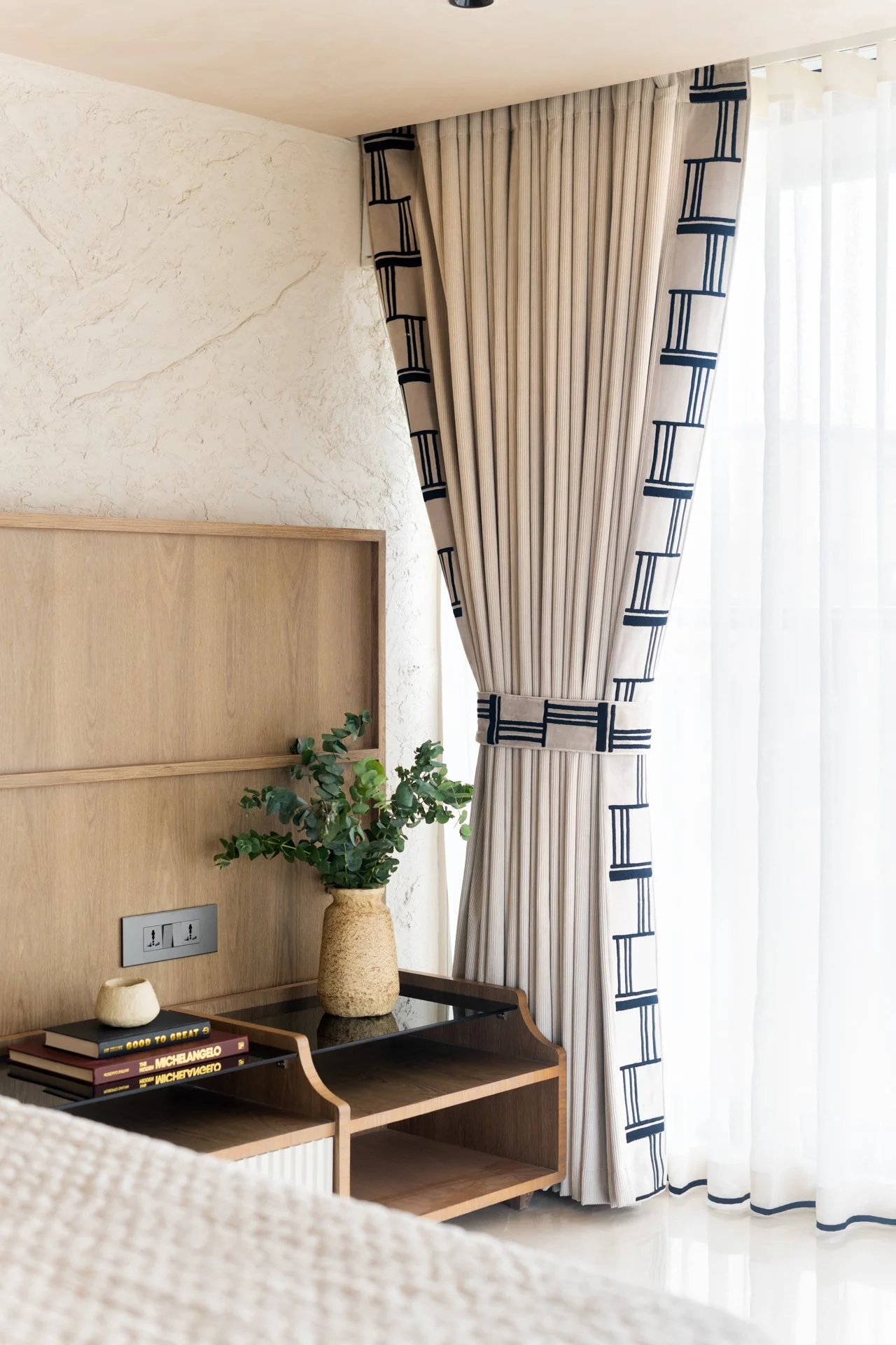 A room corner featuring a beige curtain with a black geometric border, a textured cream wall, a wooden headboard, a black tray table with books and decorative objects, a potted green plant, and a window with sheer white curtains.