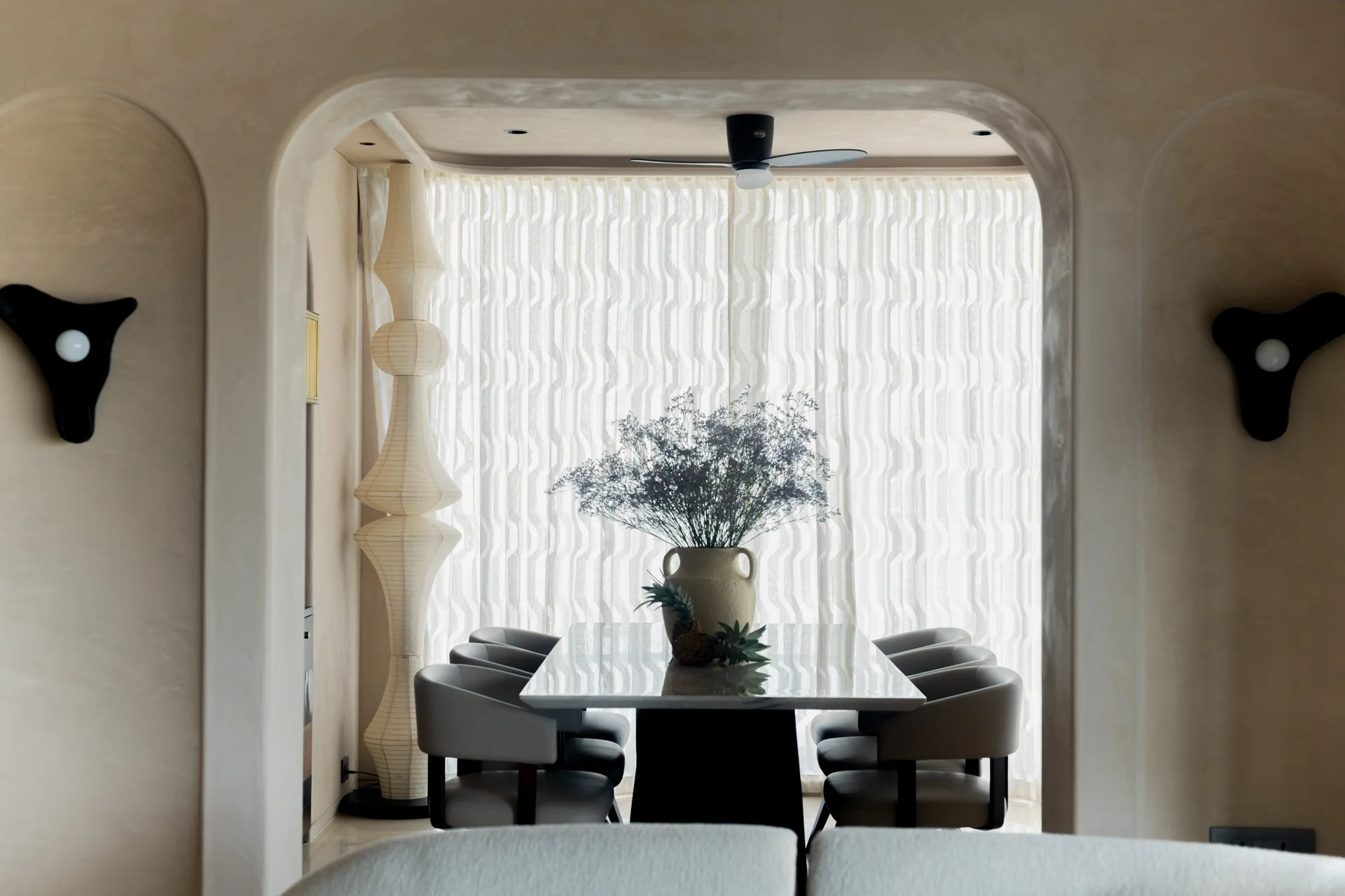 Dining room with a table and six chairs, modern decor, a large vase with greenery, and a sheer curtain letting in natural light.