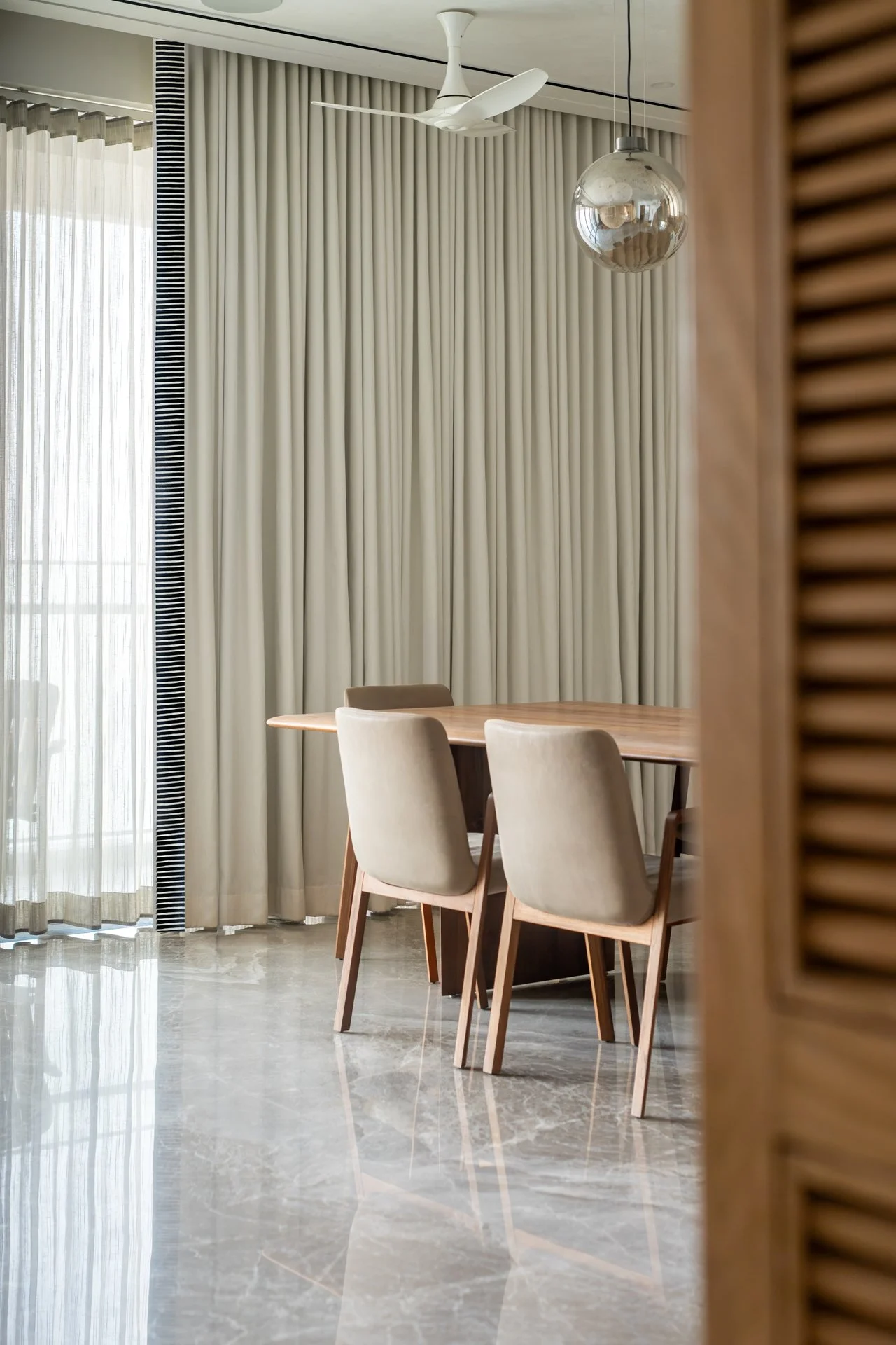 A dining room with a wooden table, four beige upholstered chairs, beige curtains, a glass pendant light, and a ceiling fan.