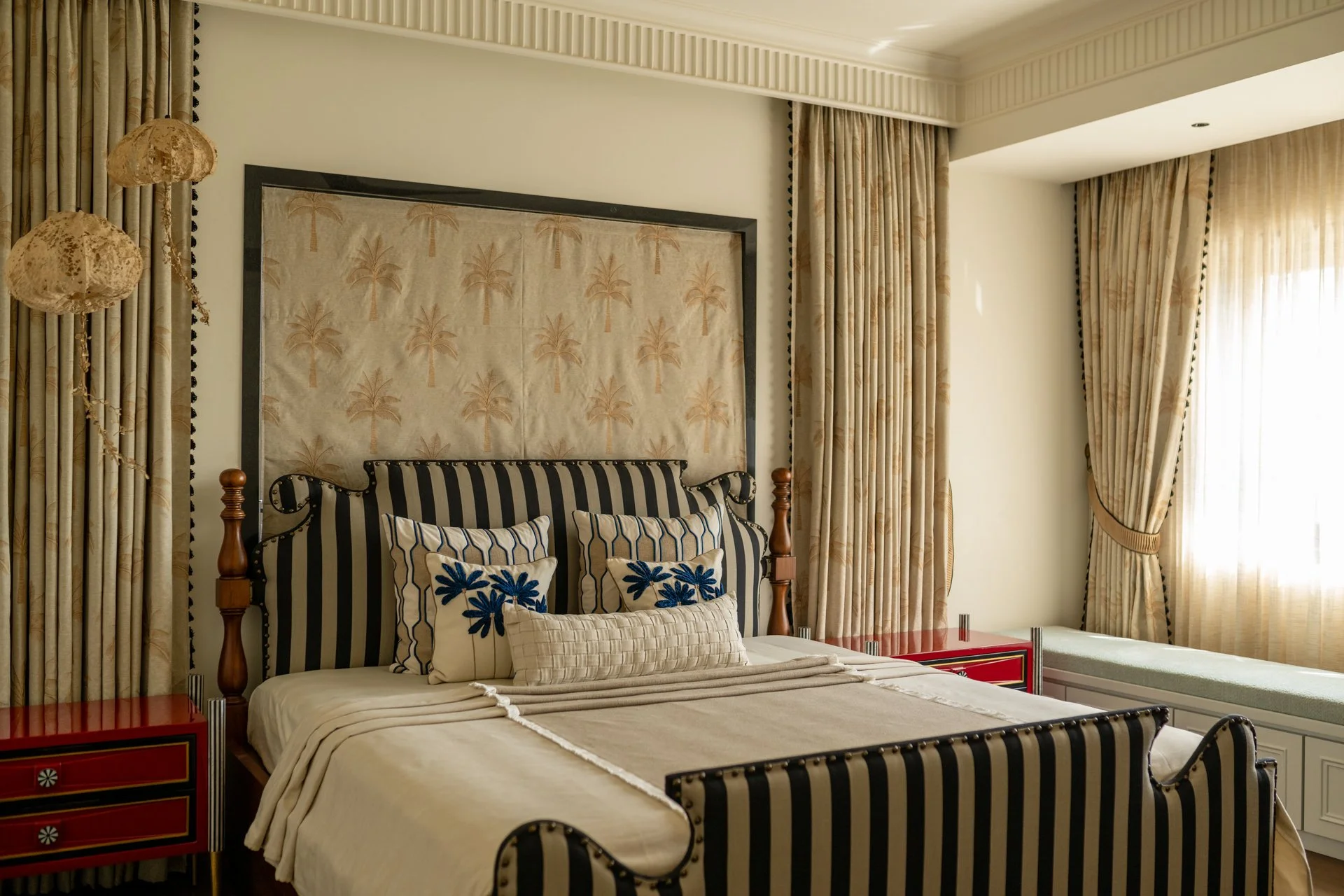 A bedroom with beige walls, a bed with a striped black and beige headboard, decorated with pillows featuring blue floral designs, flanked by cream-colored curtains with palm tree patterns, and a crystal chandelier lamp hanging on the left.