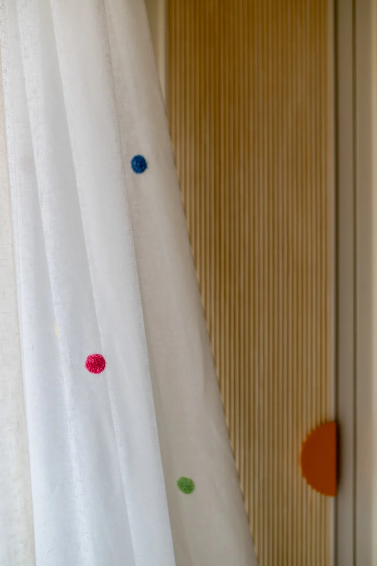 Close-up of a white curtain with colored dots, hanging beside a brown, textured wall and a wooden surface with a half-visible orange circle.