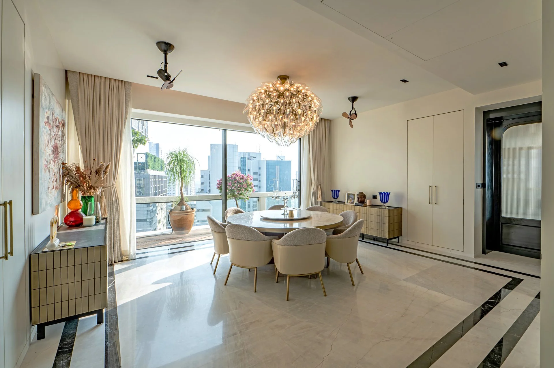 Modern dining room with round table surrounded by beige chairs, large window with city view, decorative vases, and chandelier, with a sideboard and art on the wall.