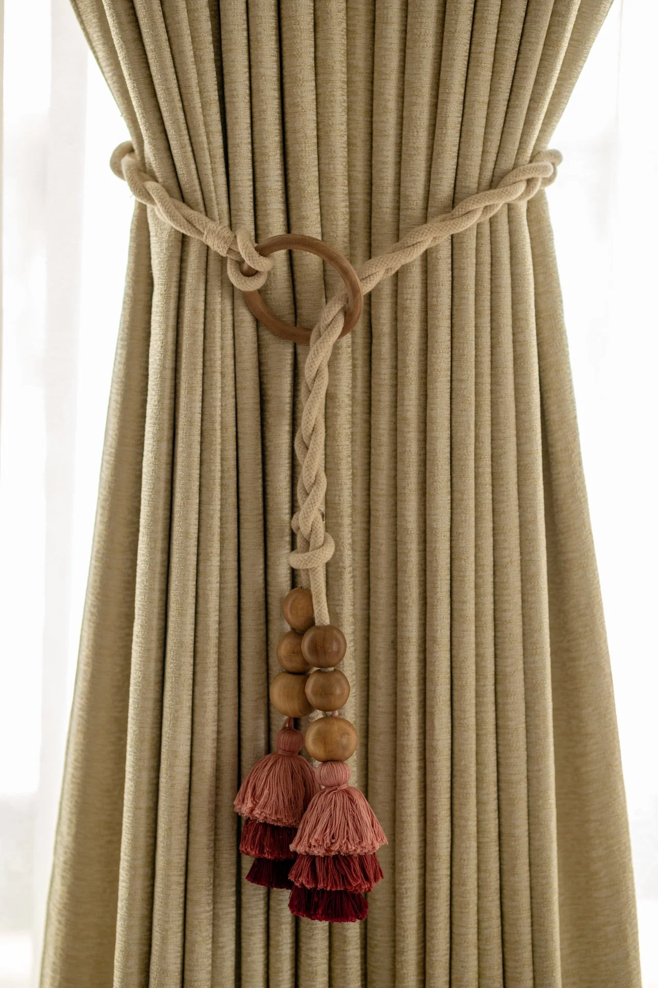 Beige curtains held back with a beige braided rope tieback featuring large wooden beads and pink tassels.