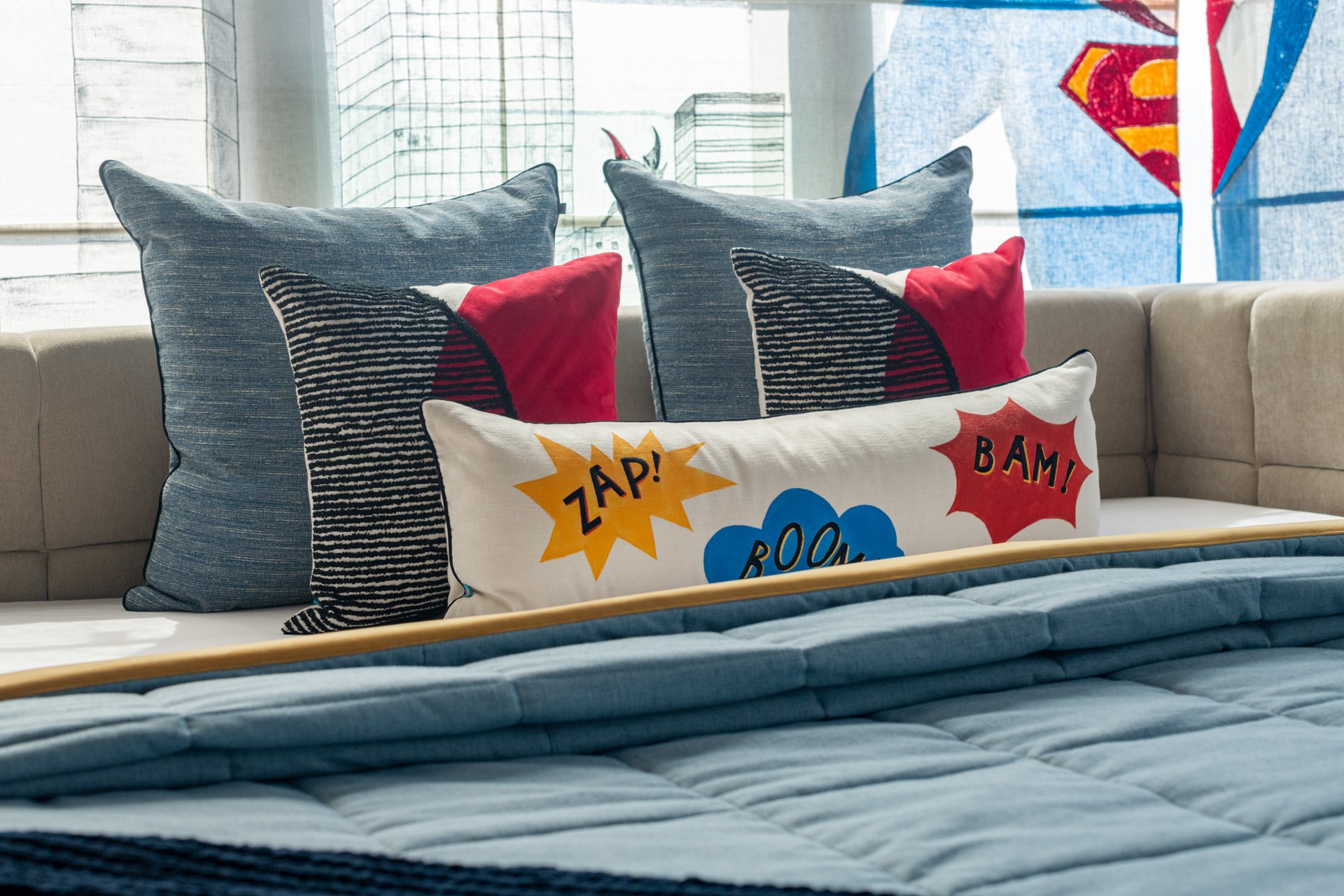 Close-up of decorative pillows on a beige couch, including one with comic book style words like 'ZAP!', 'BOOM!', and 'BAM!' in bright colors, with cityscape and superhero logos in the background.