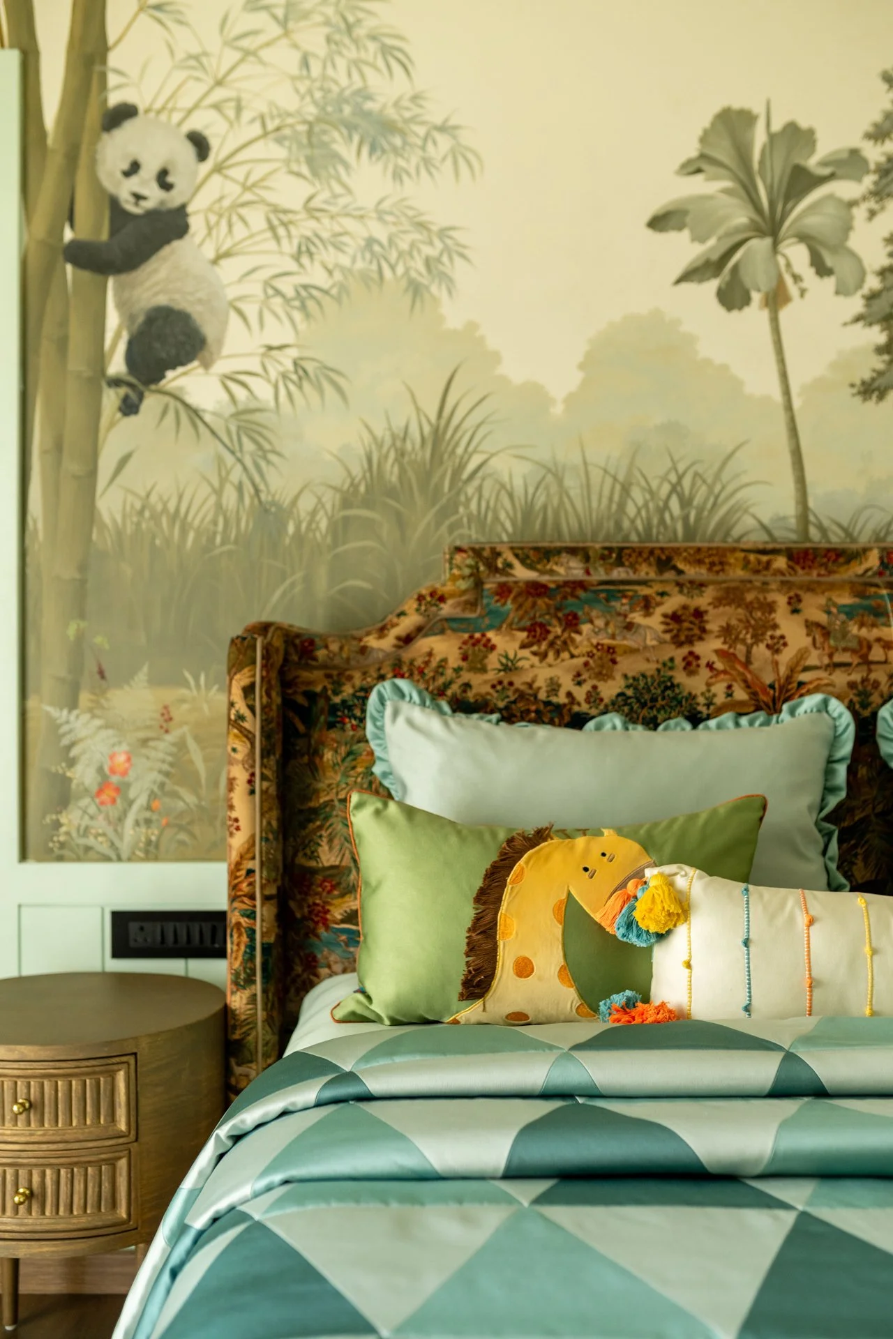 Decorative bedroom wall with panda mural, floral wallpaper, colorful pillows on a bed with a diamond pattern bedspread, and a small wooden bedside table.
