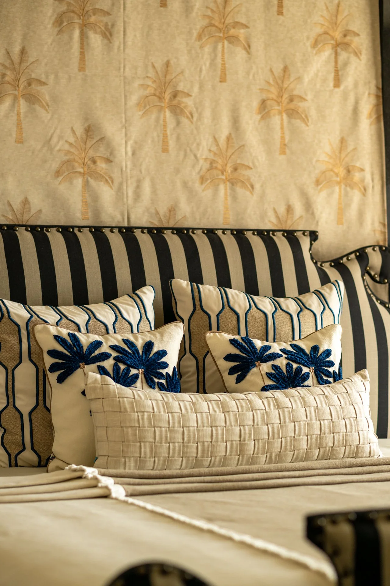 A bed with a striped black and cream headboard and decorative pillows, including two with blue embroidered floral designs and a woven beige pillow in front. There is beige bedding and a bedspread in the foreground.