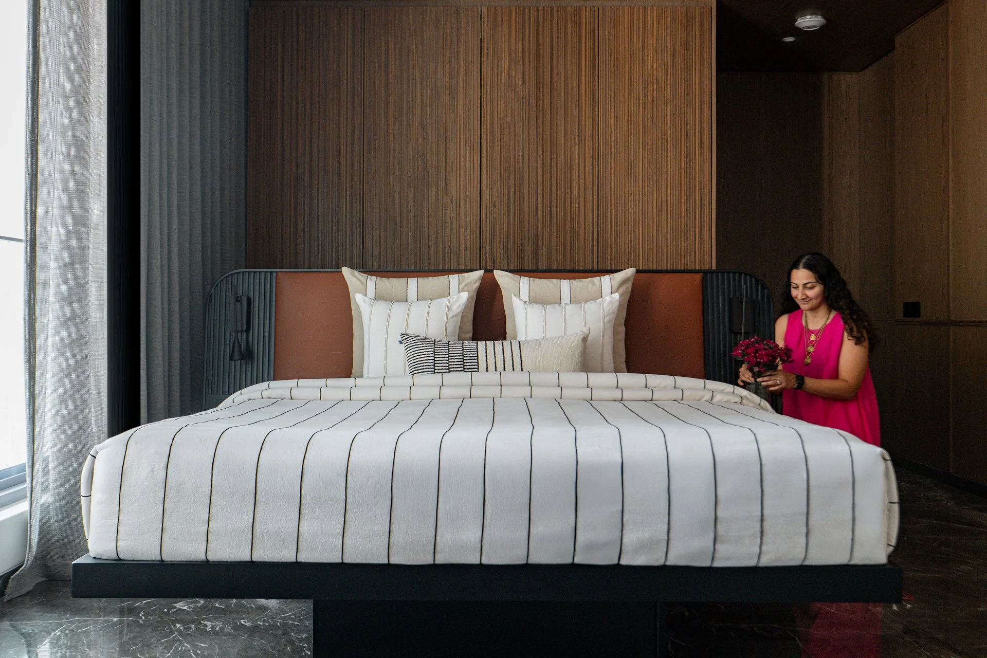A bedroom with a large bed with white bedding with black stripes, decorated with multiple pillows. A woman with dark curly hair wearing a pink dress, holding a flower bouquet, stands beside the bed. The room has wood-paneled walls, dark curtains, and marble flooring.