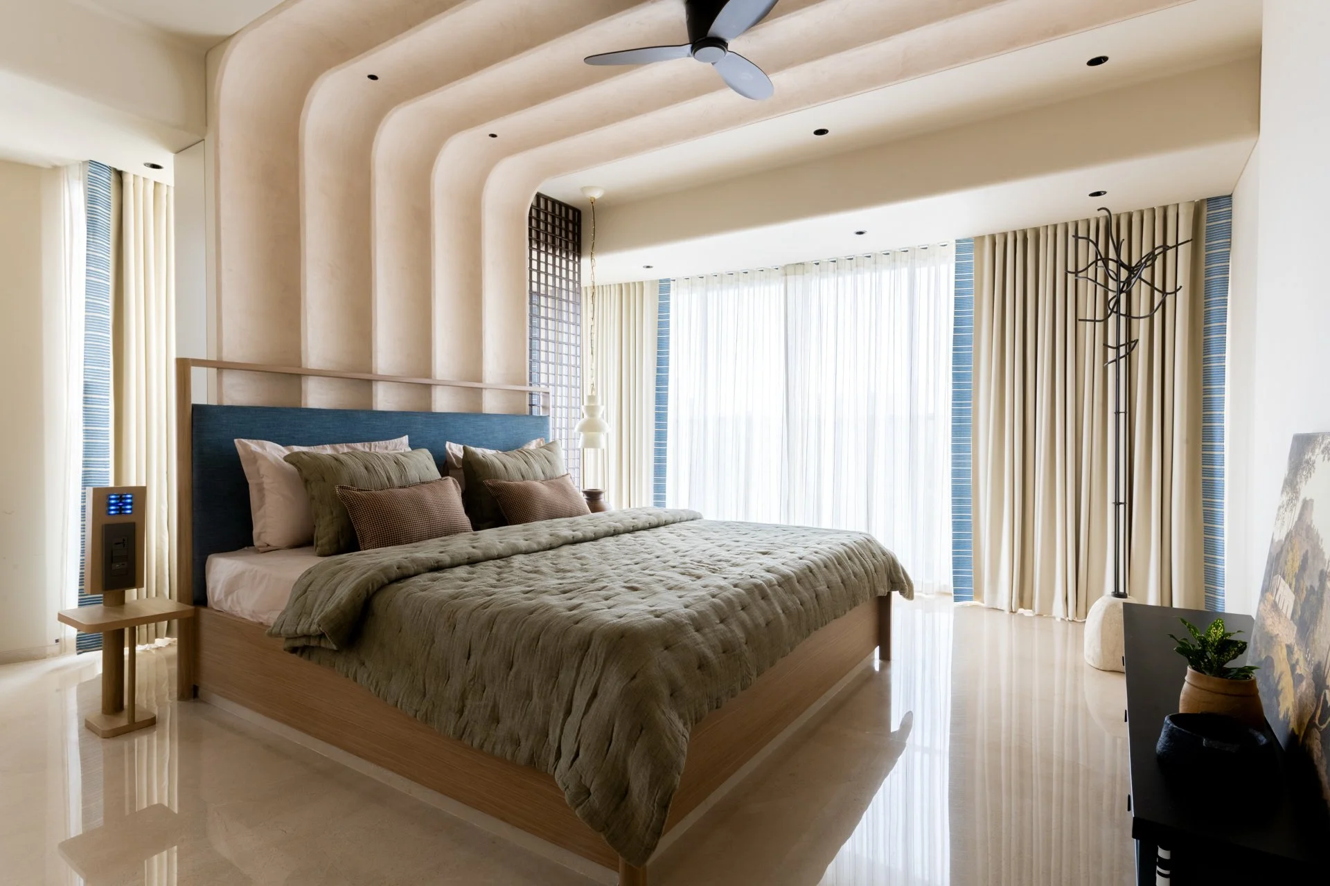 Modern bedroom with large bed, beige and green bedding, and decorative pillows. Floor-to-ceiling windows with sheer curtains and cream drapes. Ceiling fan and unique floor lamp. Light-colored flooring and minimalist decor.